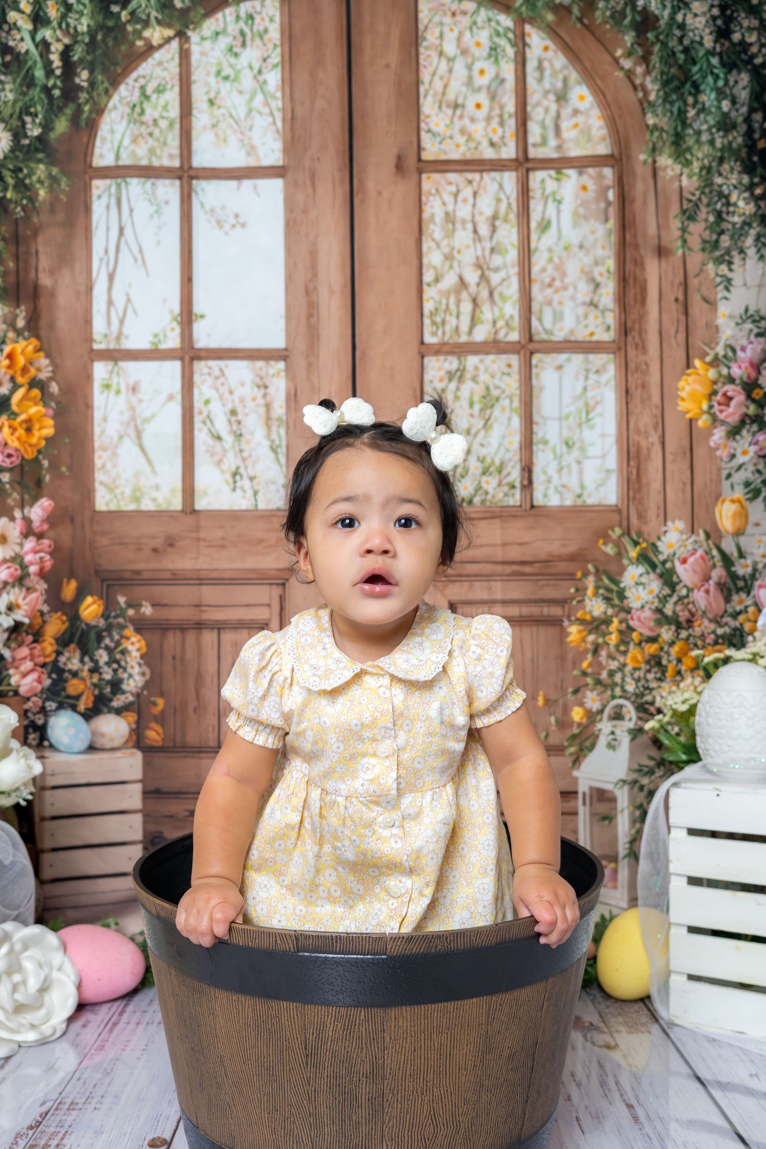 Toddler girl in yellow floral dress sitting in wooden barrel tub with Easter eggs and floral backdrop.