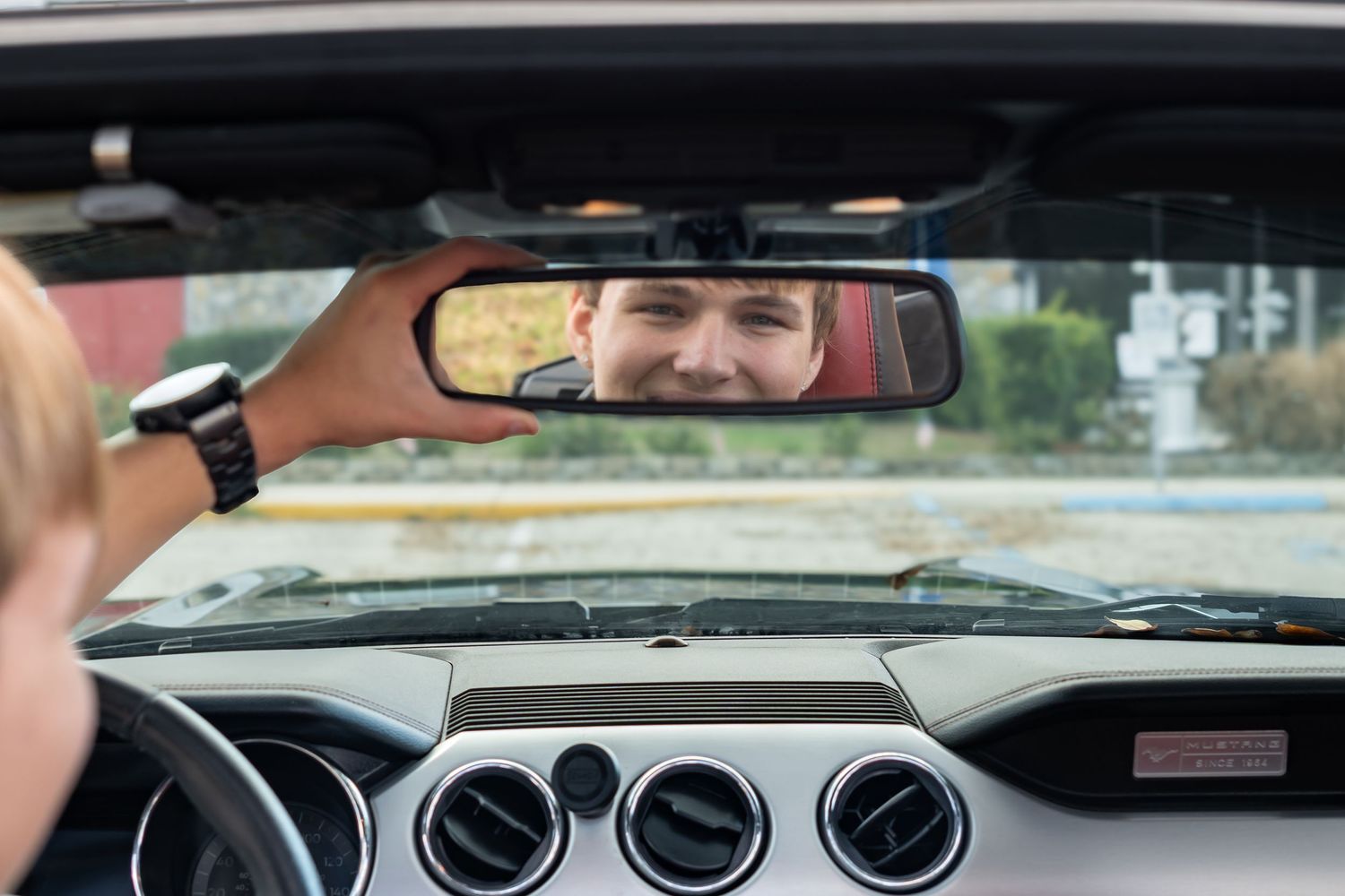 Man smiling in rearview mirror of convertible car while driving on a sunny day.