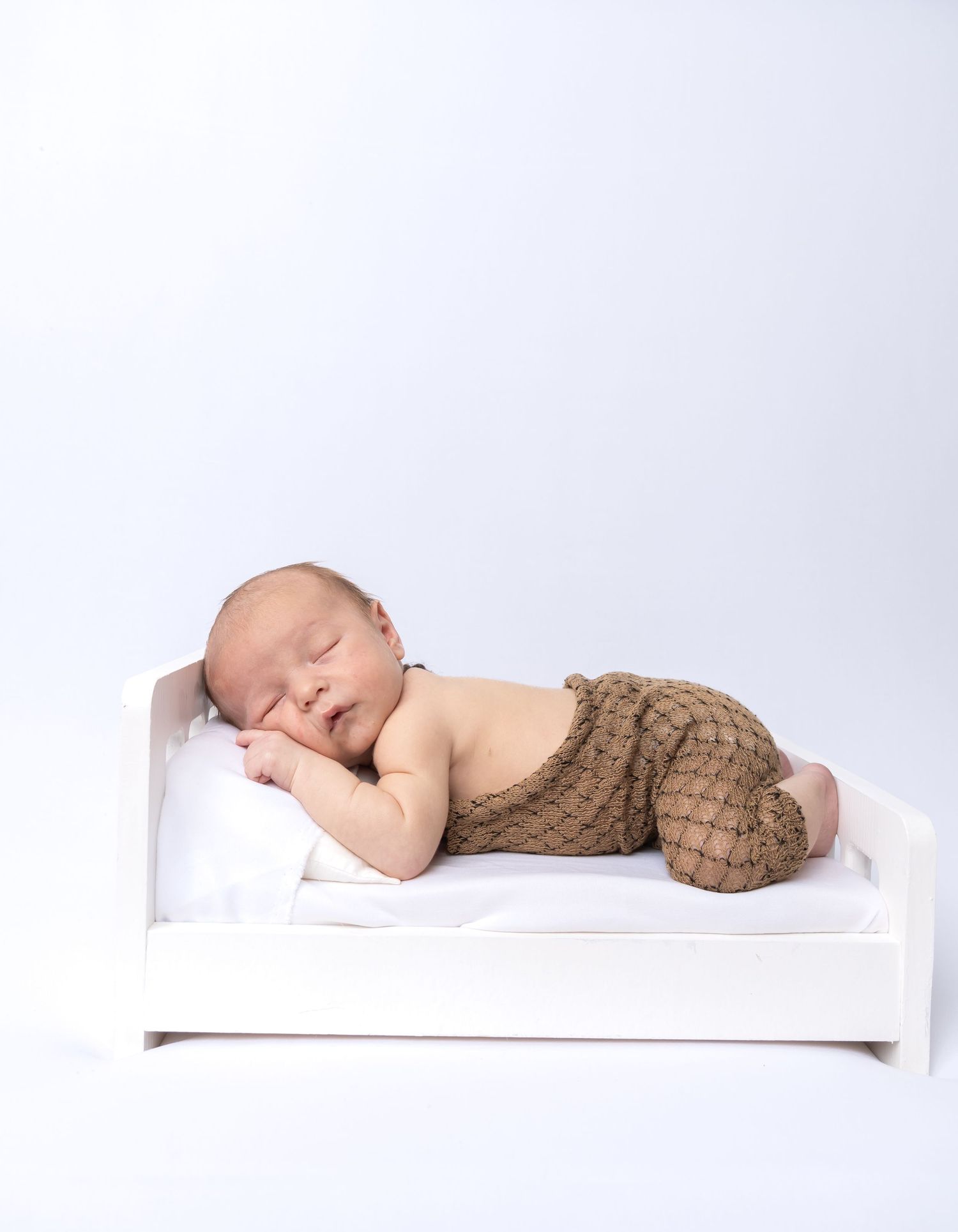 Newborn baby sleeping peacefully on a white miniature bed, wearing brown knit pants, on a light background.