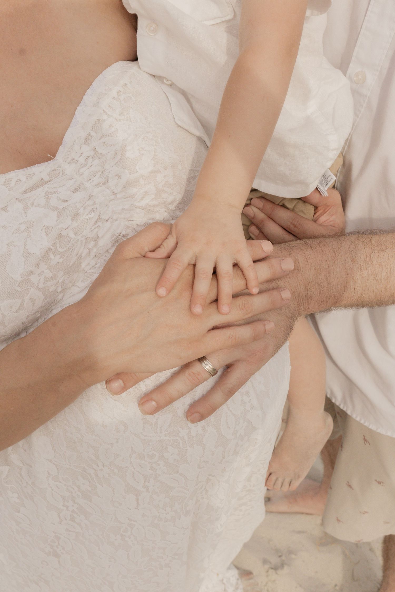 Close-up of family hands resting on pregnant womans belly, showing wedding ring on mothers hand at beach.