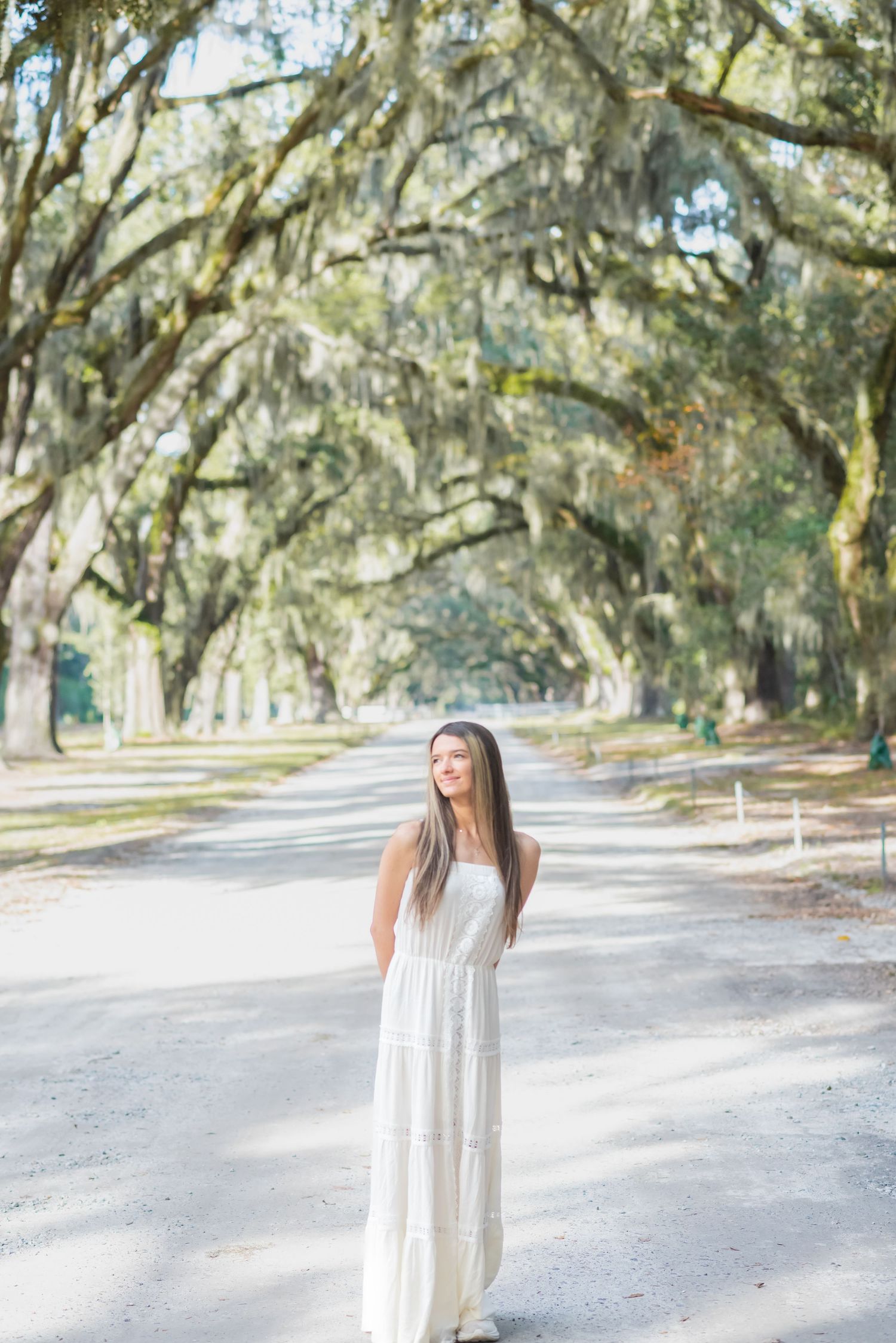Young woman in white maxi dress standing on a tree-lined path with Spanish moss-draped oak trees.