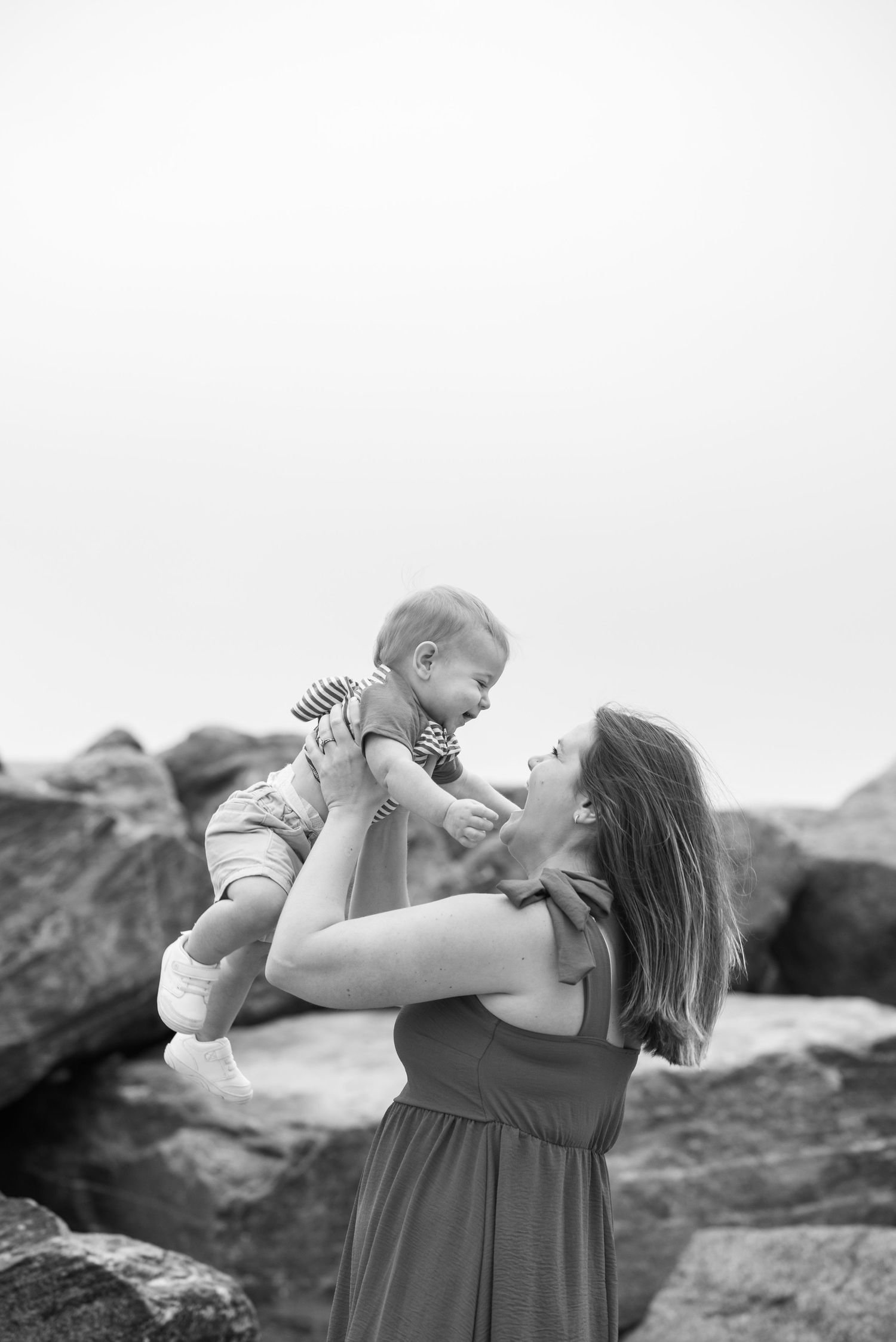 Mother joyfully lifting laughing baby in black and white beach photo.