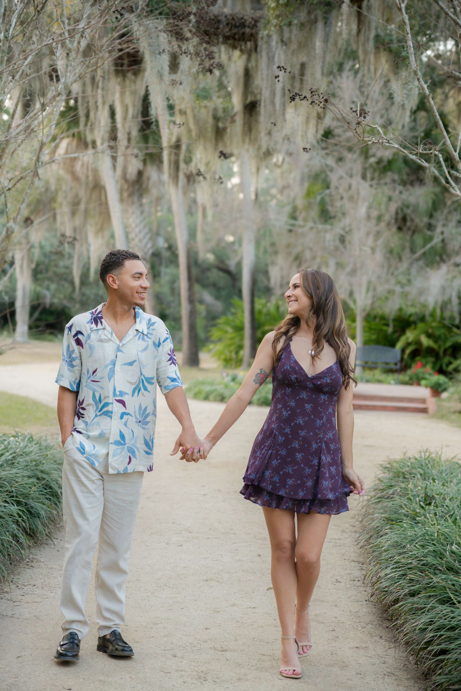 A smiling couple holds hands walking on a garden path, man in floral shirt and woman in purple mini dress.