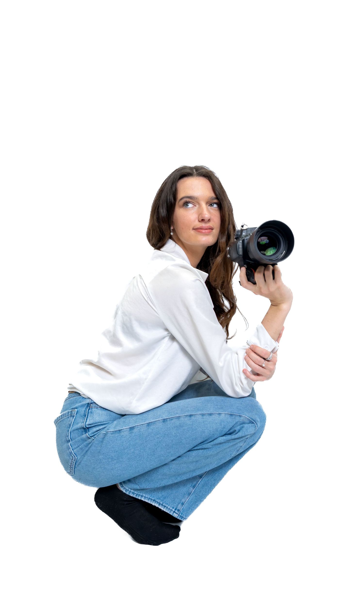 Female photographer crouching with a professional DSLR camera, wearing white blouse and jeans on white background.