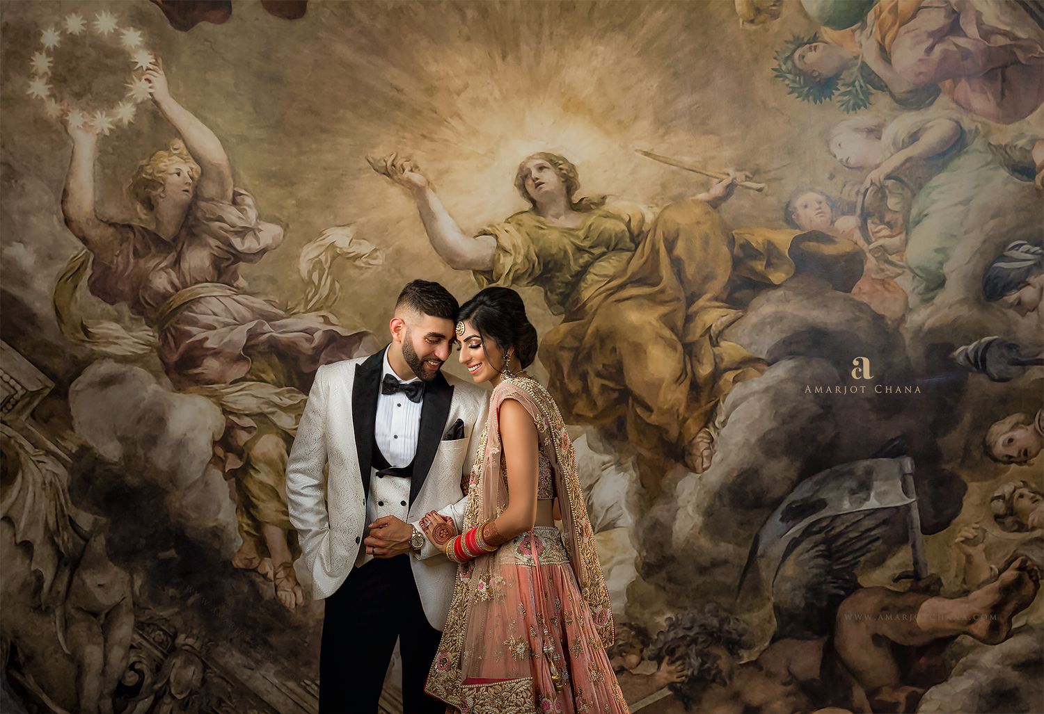 Indian wedding couple in bridal attire pose before stunning Renaissance-style painted mural.