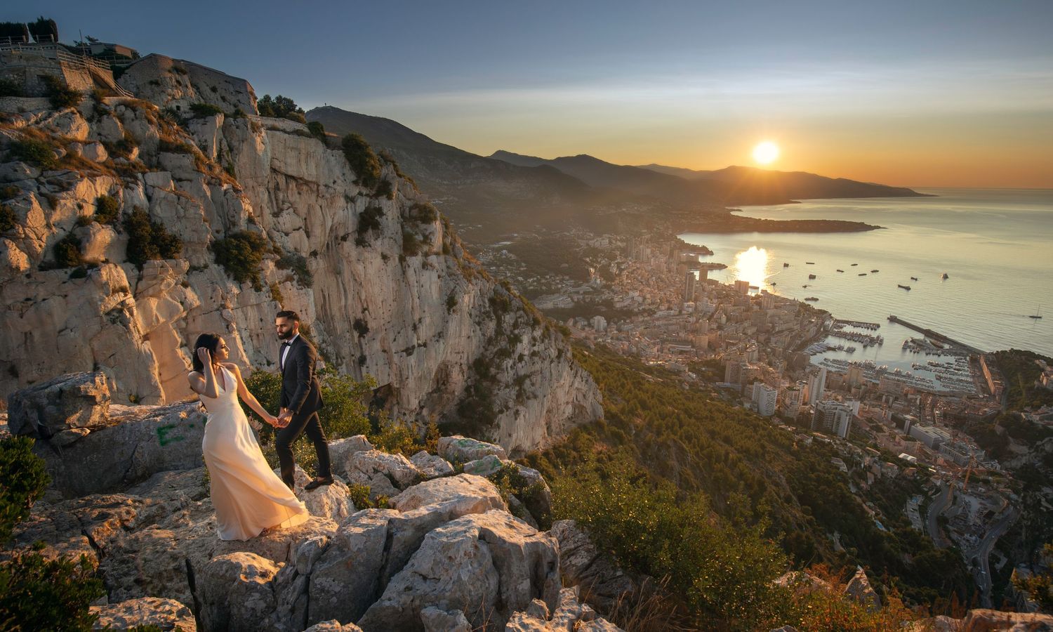 Couple holding hands on rocky cliffside overlooking coastal city at golden sunset.