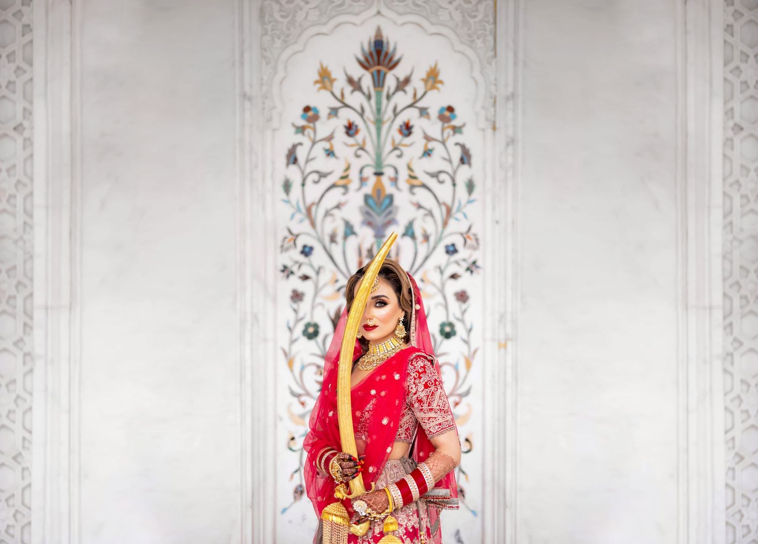 Indian bride in red bridal lehenga posing gracefully before ornate marble floral wall inlay at historic Indian monument, wedding photography