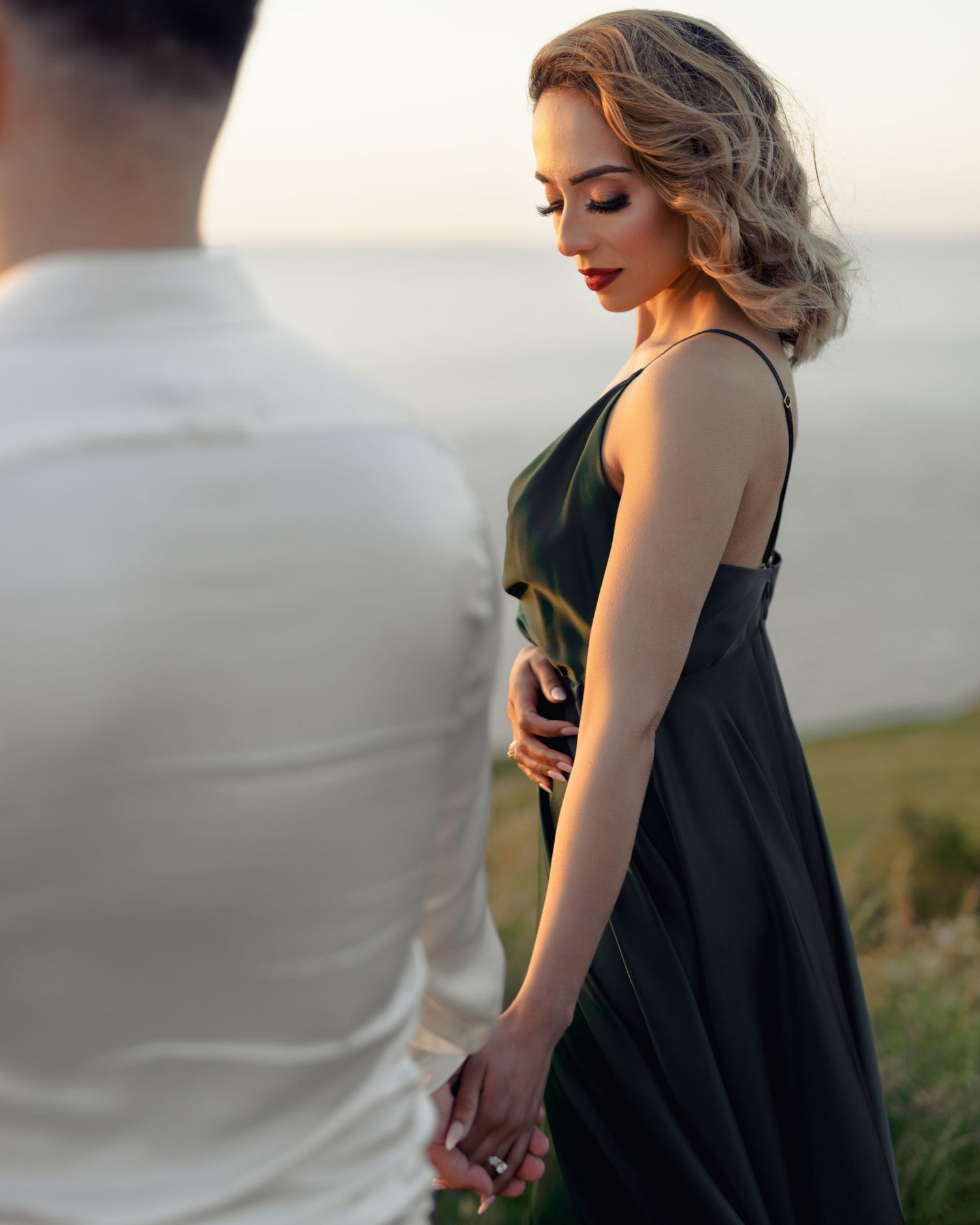 A couple holds hands outdoors at sunset, woman in elegant green dress with red lips, man in white shirt.