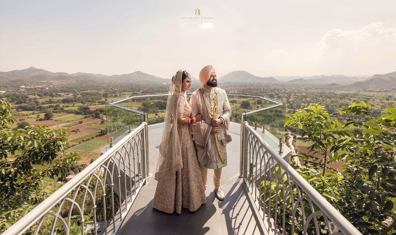 Indian bride and groom in traditional attire on a scenic bridge overlooking lush green valley and mountains.