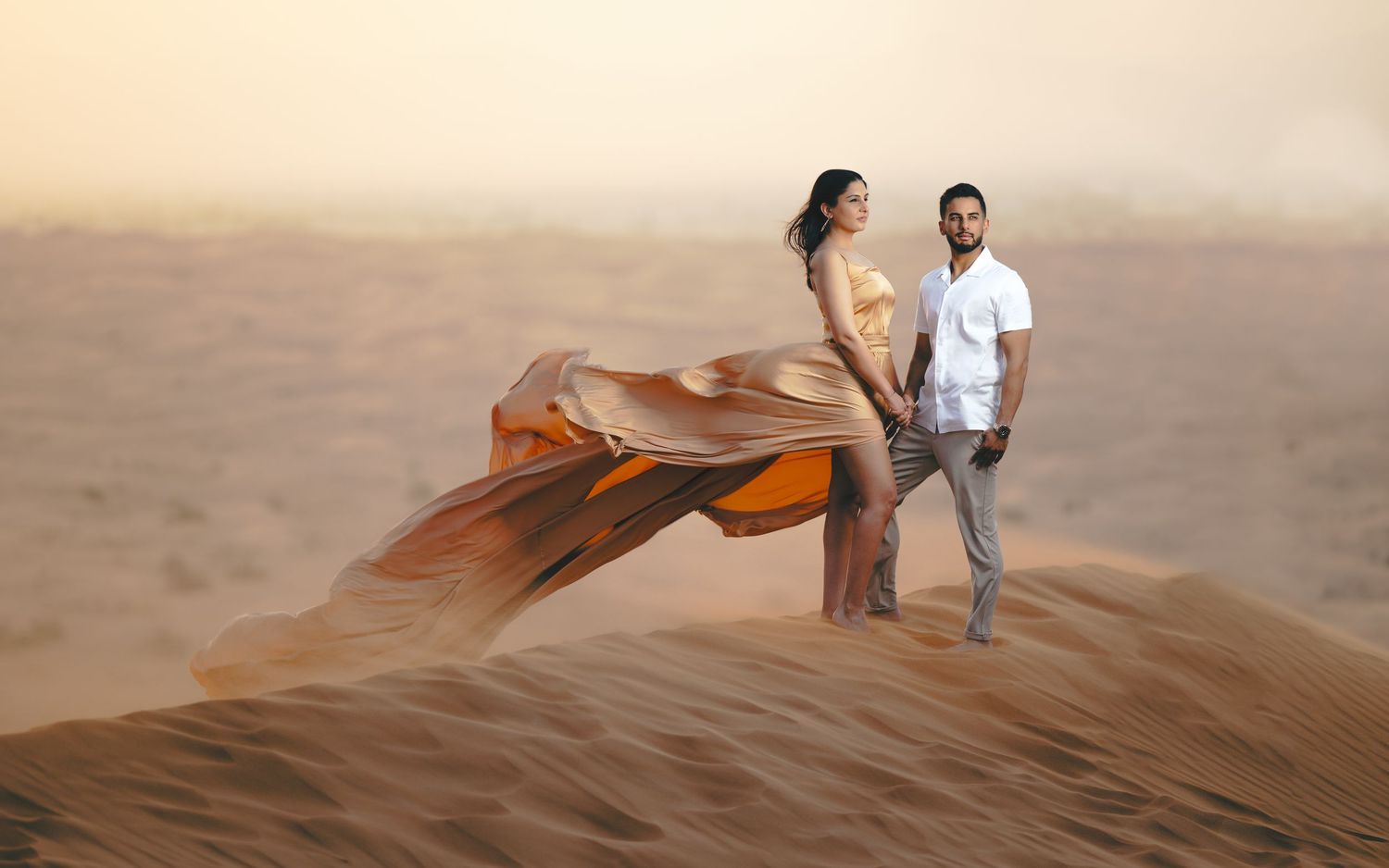 Sam and Sapna pre-wedding portraits – couple posing dramatically on desert sand dunes, woman in flowing orange gown billowing in wind, desti