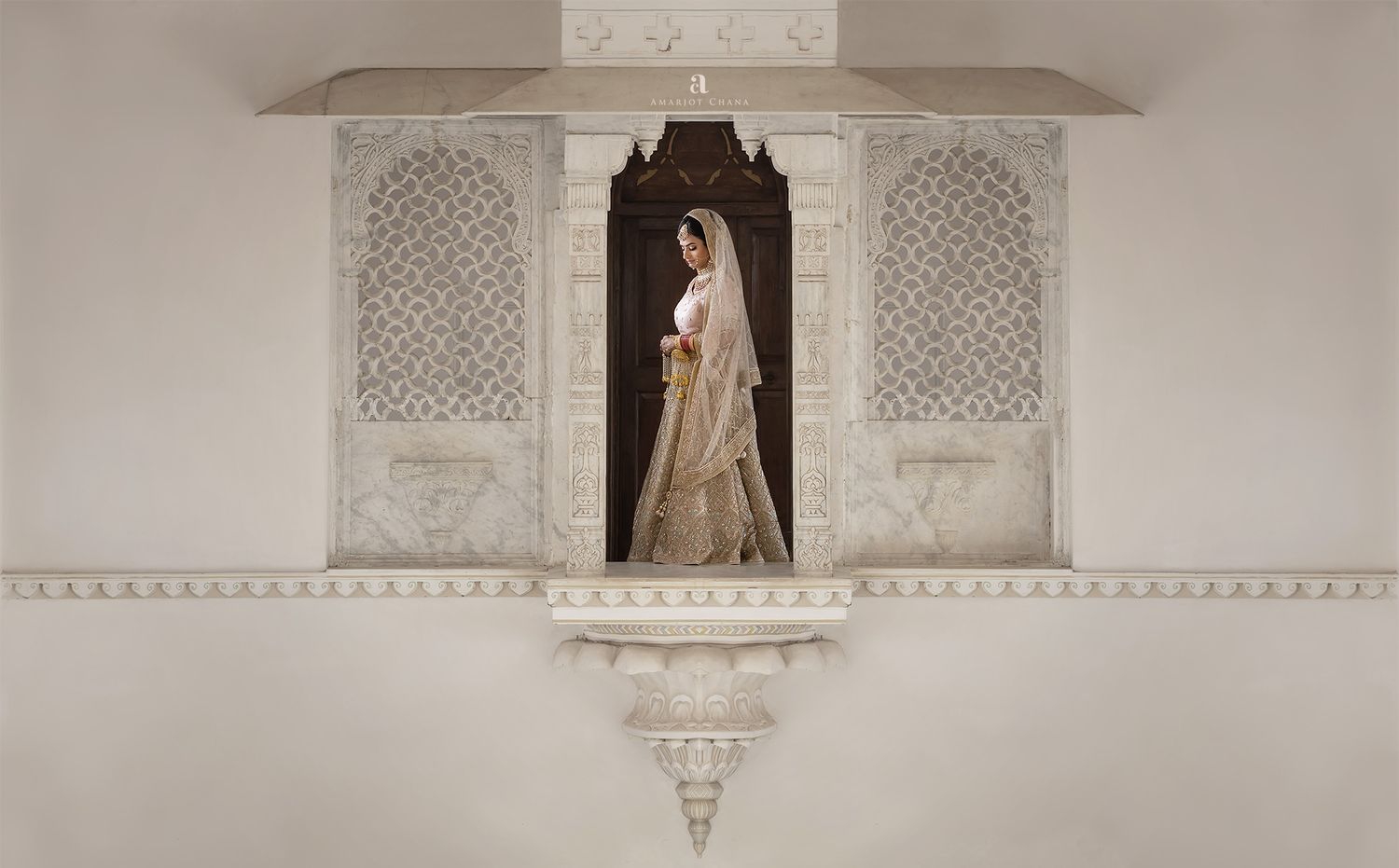 A bride in traditional Indian bridal attire stands in an ornate white marble archway of a palace.