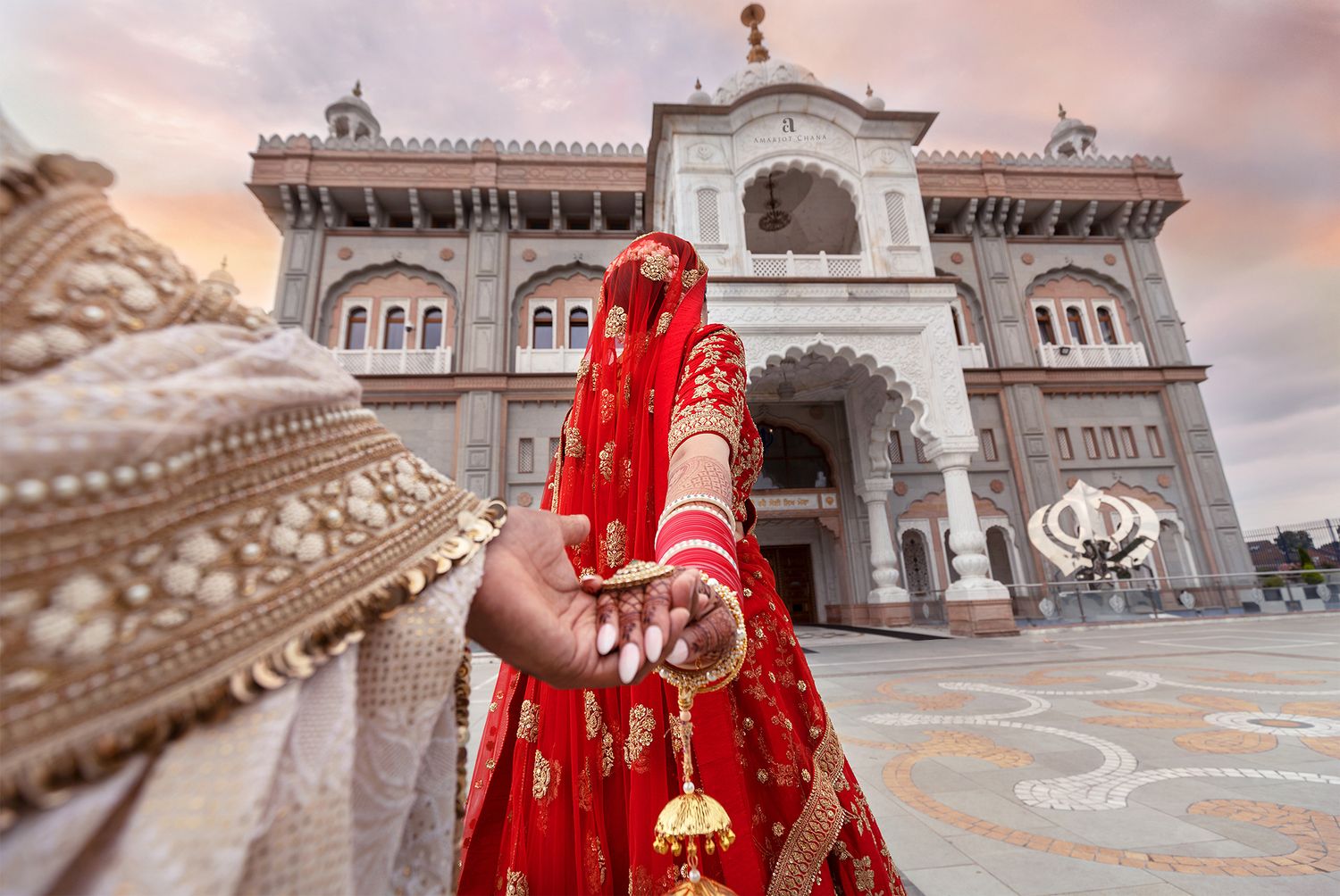 Indian bride in red lehenga holds groom's hand outside ornate Sikh temple at sunset wedding ceremony.
