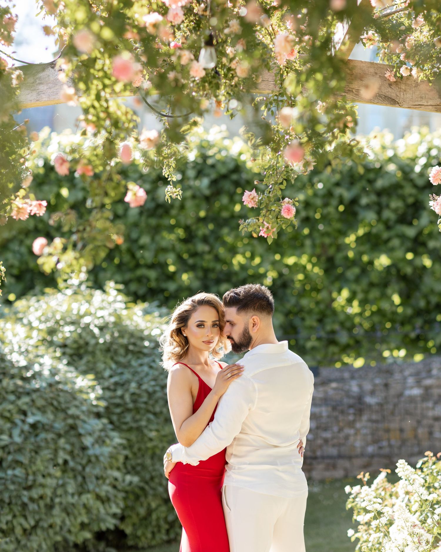 Inda and Sandie pre-wedding shoot – couple embracing in romantic pink rose garden, woman in red dress, outdoor Indian wedding portrait photo