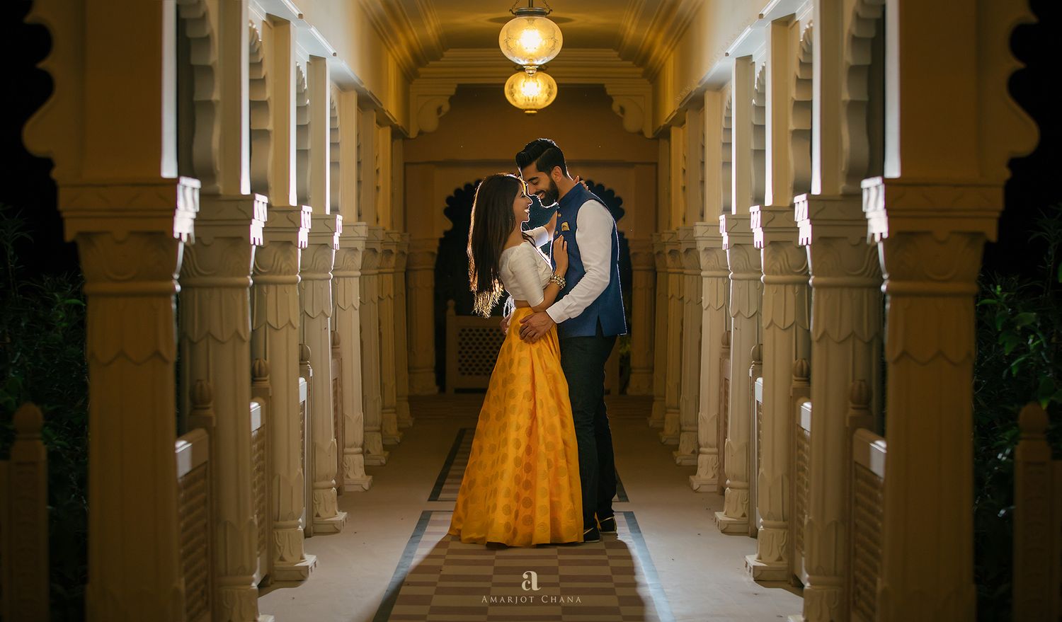 Romantic couple embracing in an ornate golden corridor, woman in yellow lehenga and man in blue jacket.