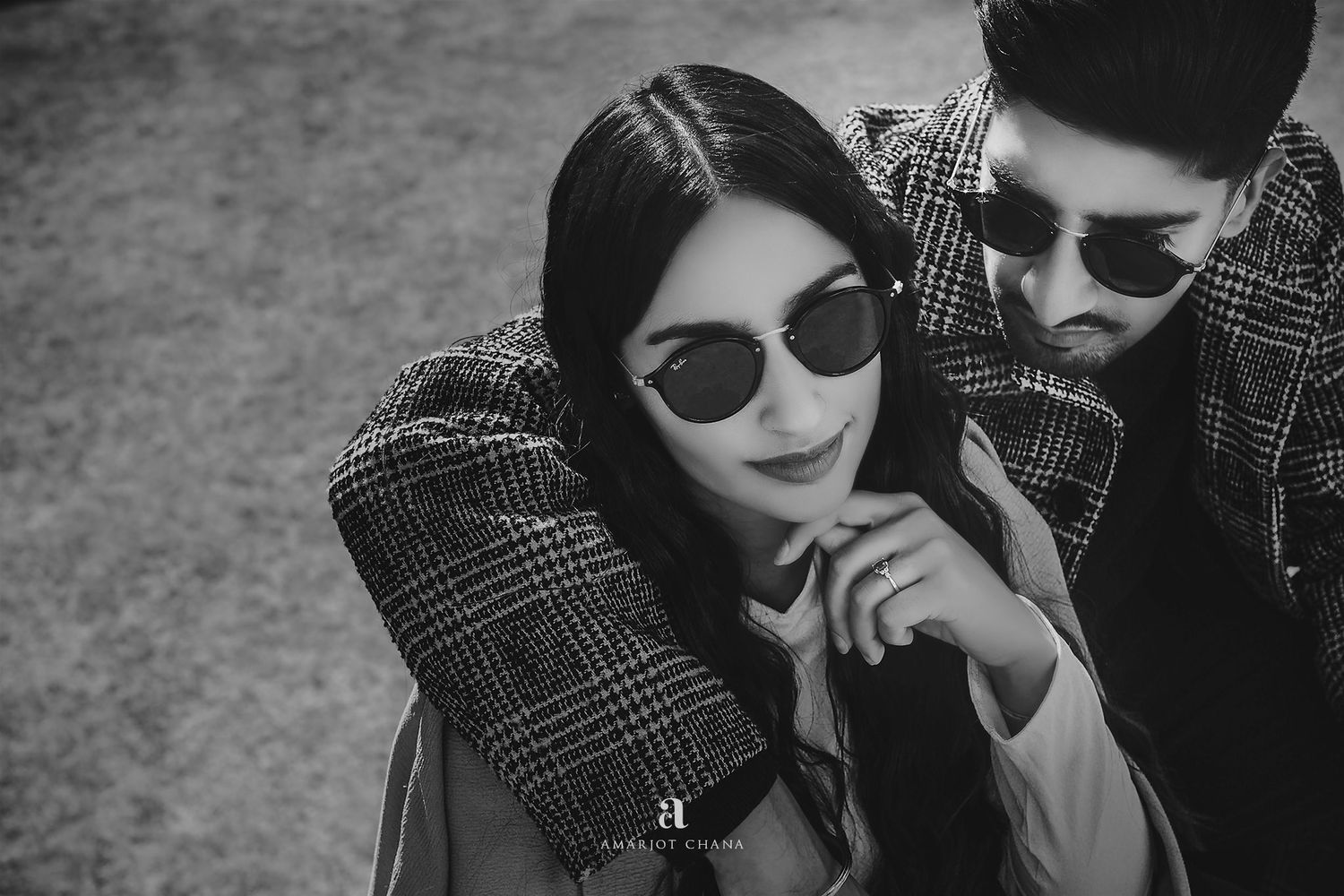 Black and white photo of a stylish couple wearing sunglasses and plaid coats, posing closely together outdoors.