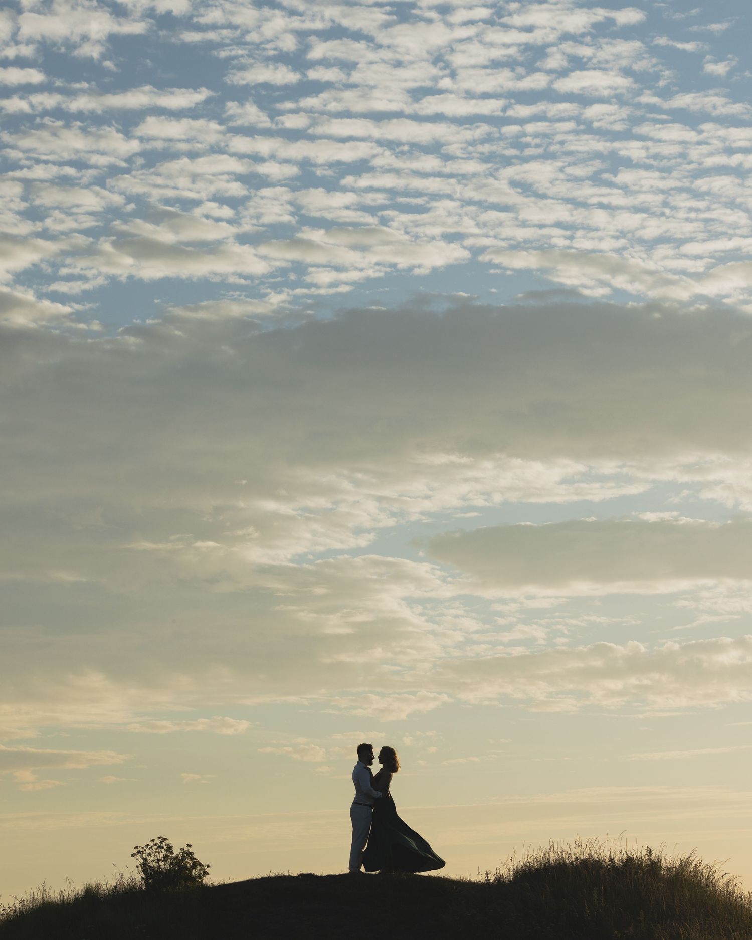 Pre-wedding shoot – couple silhouette embracing on hilltop beneath dramatic cloudy sky at golden sunset, Indian wedding photography