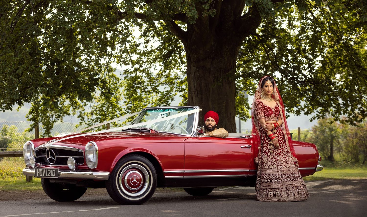 Sikh couple in traditional red wedding attire posing beside a classic red Mercedes-Benz convertible under a large tree.