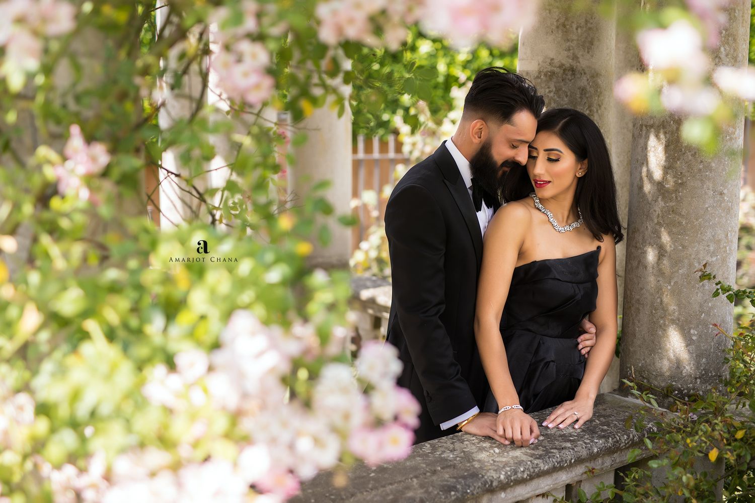 Pre-wedding shoot – couple in elegant black outfits sharing tender moment among blooming pink flowers, romantic Indian wedding photo