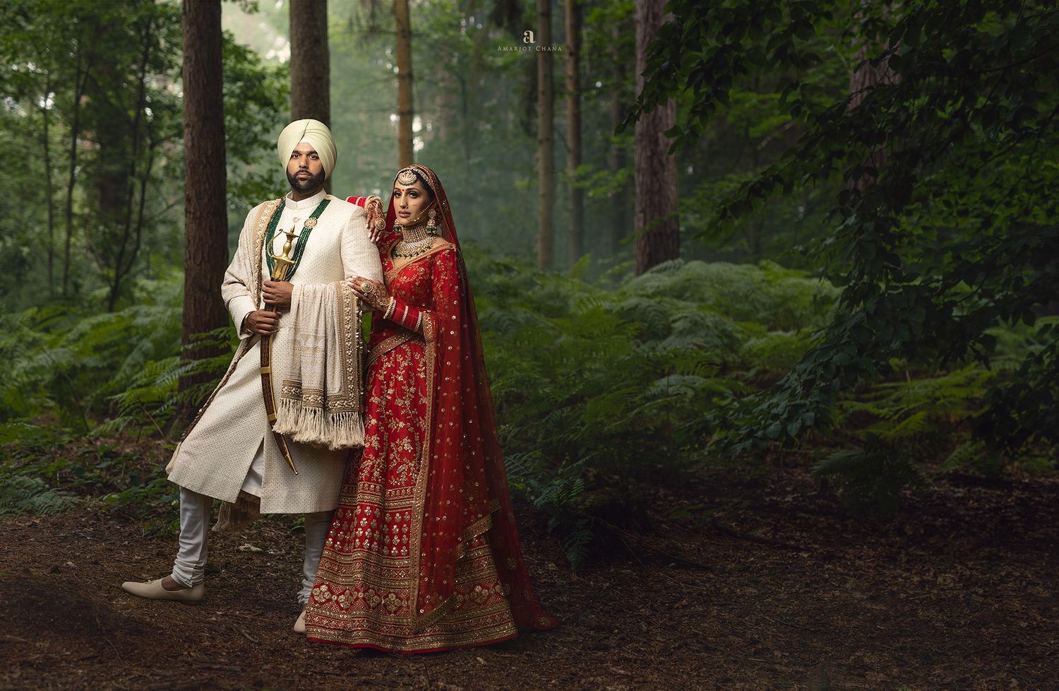 Indian bride in red lehenga and groom in white sherwani pose elegantly in a lush forest setting.