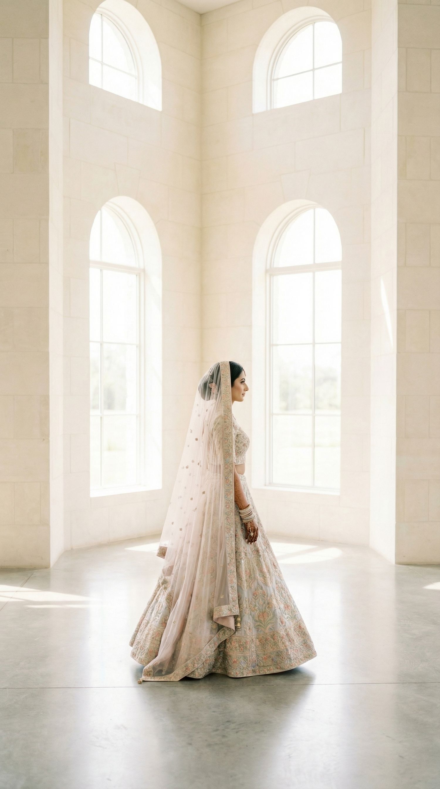 Indian bride in pastel floral lehenga with dupatta walking through bright white arched wedding venue, South Asian wedding photography UK