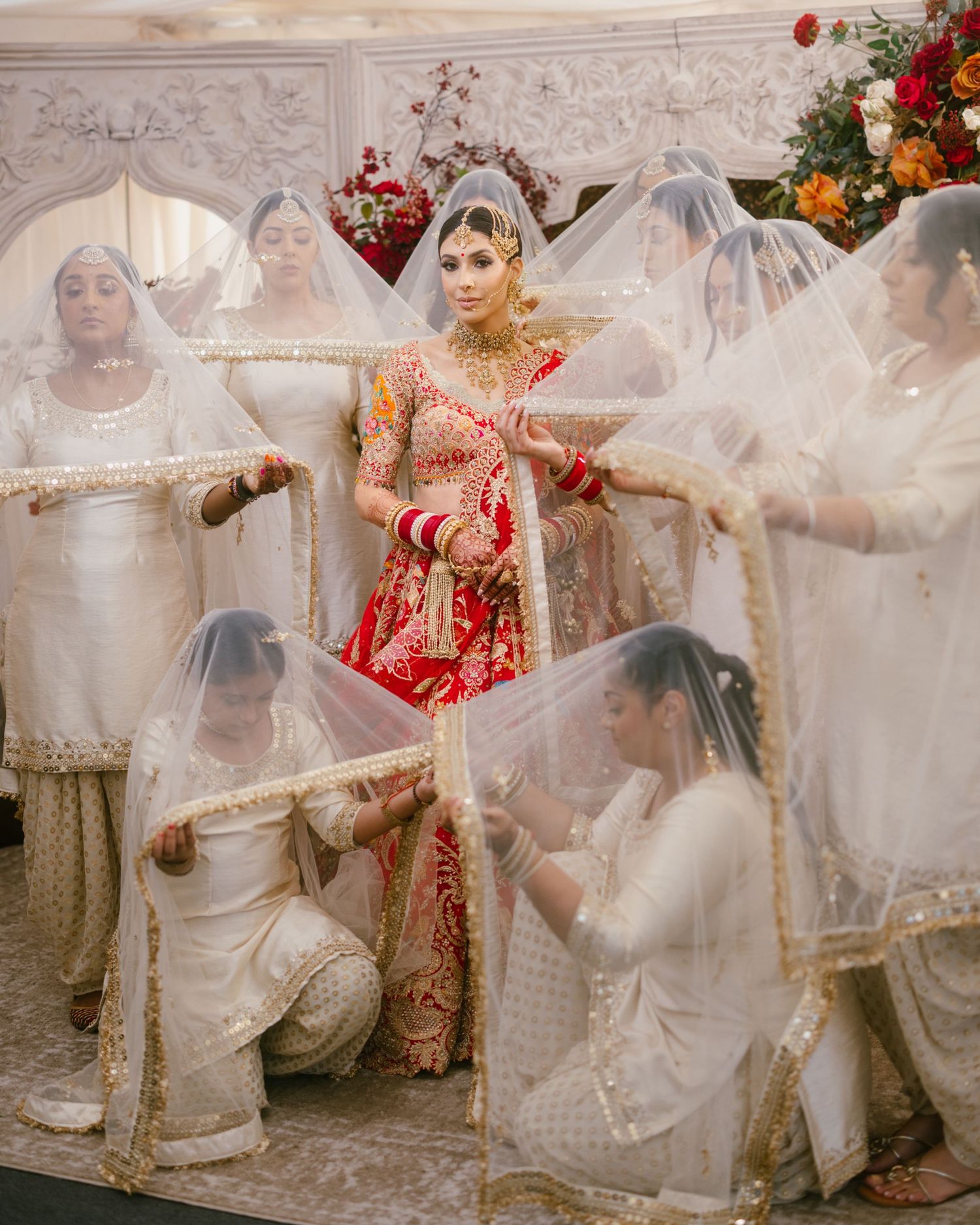 Sikh bride in red lehenga surrounded by women in white dupattas during traditional wedding ceremony.