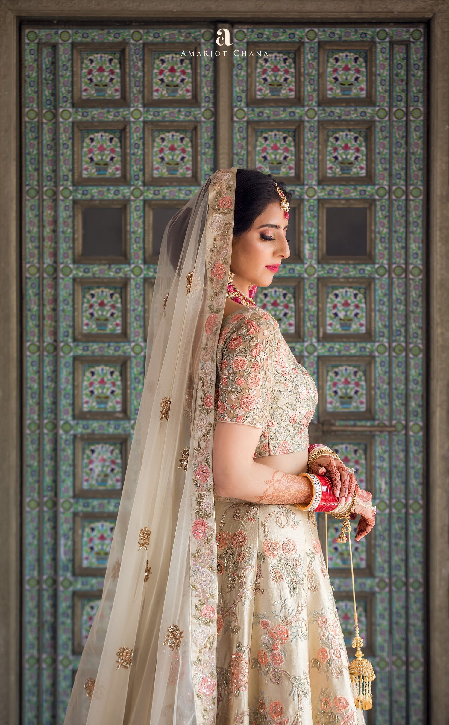 Indian bride in ivory floral lehenga with veil poses before ornate teal carved wooden doors.
