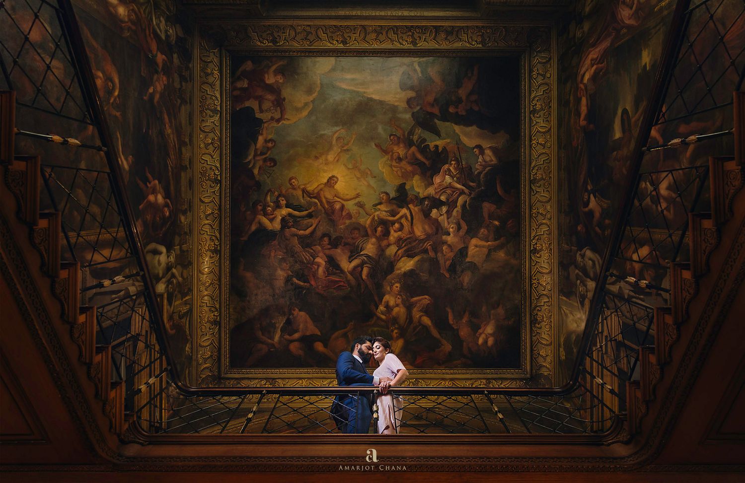 Couple sharing a romantic kiss beneath an ornate painted ceiling in a grand historic hall.