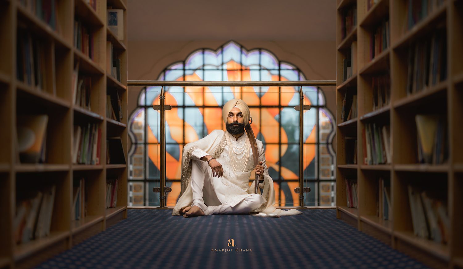Sikh man meditating in lotus pose between library bookshelves with ornate stained glass window backdrop.