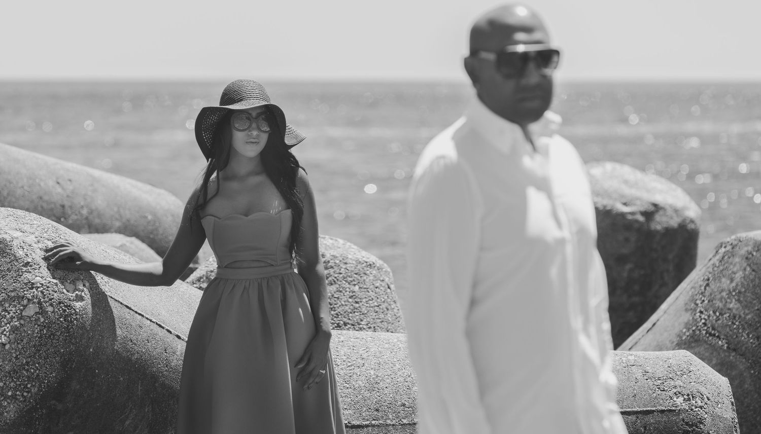 Robin and Jyoti pre-wedding portraits – artistic black and white photo of couple by rocky seaside, woman in hat and dress, dramatic coastal