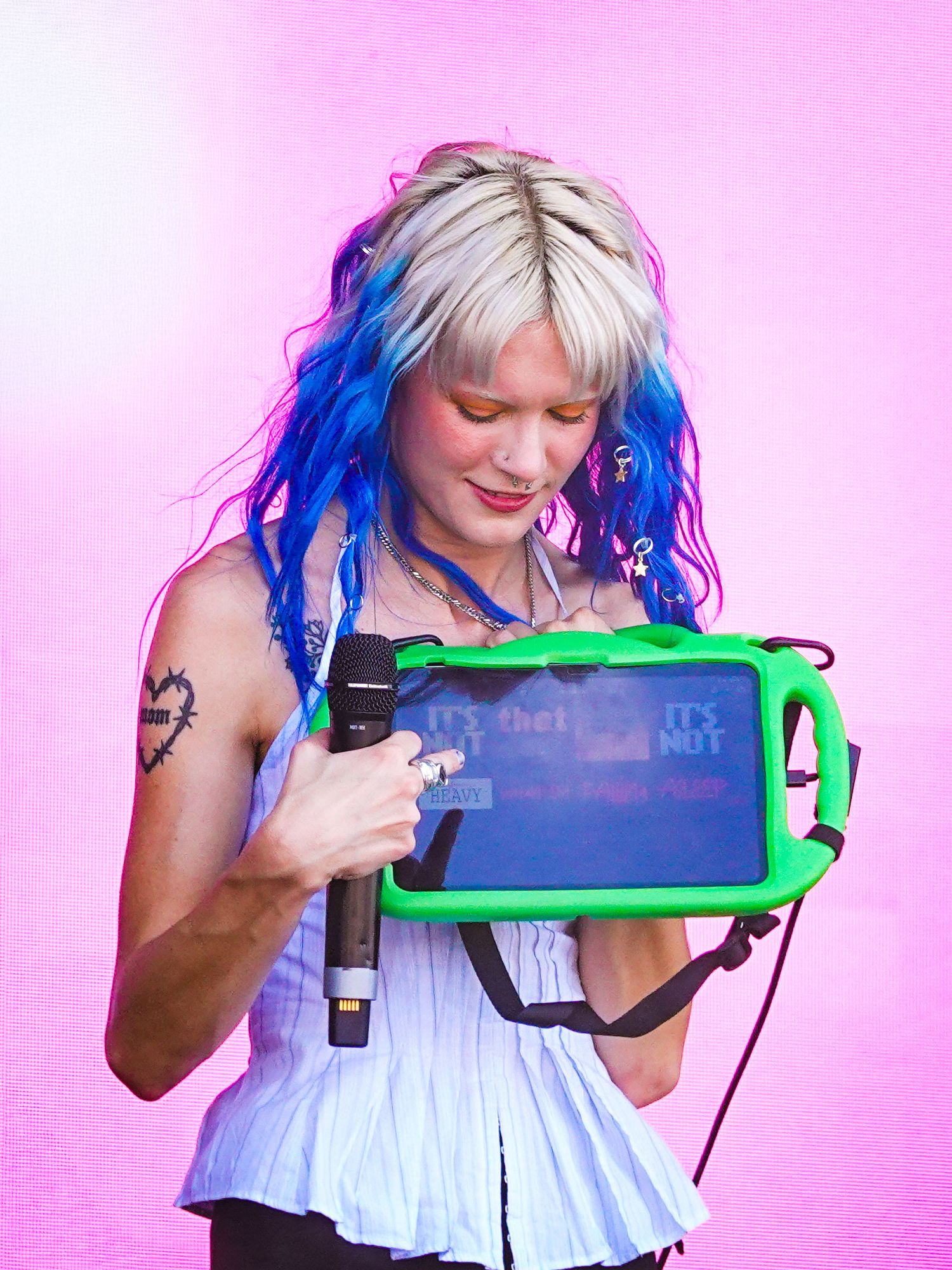Young woman with blue and white hair holds a green-cased tablet and microphone against a pink background.