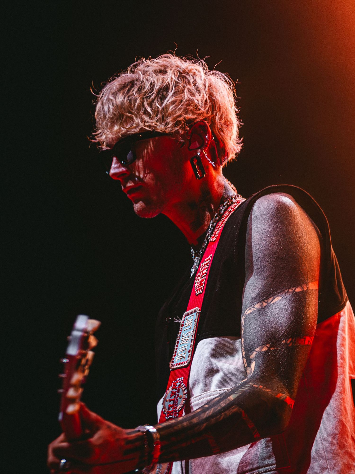 A rock musician with curly blonde hair and sunglasses plays electric guitar on stage with a red strap under dramatic lighting.