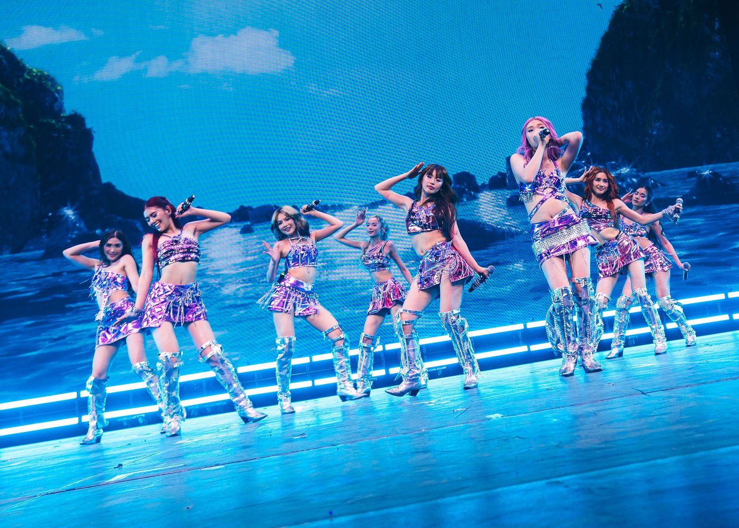 P-pop girl group performs synchronized dance routine on stage in colorful purple outfits with blue scenic backdrop.