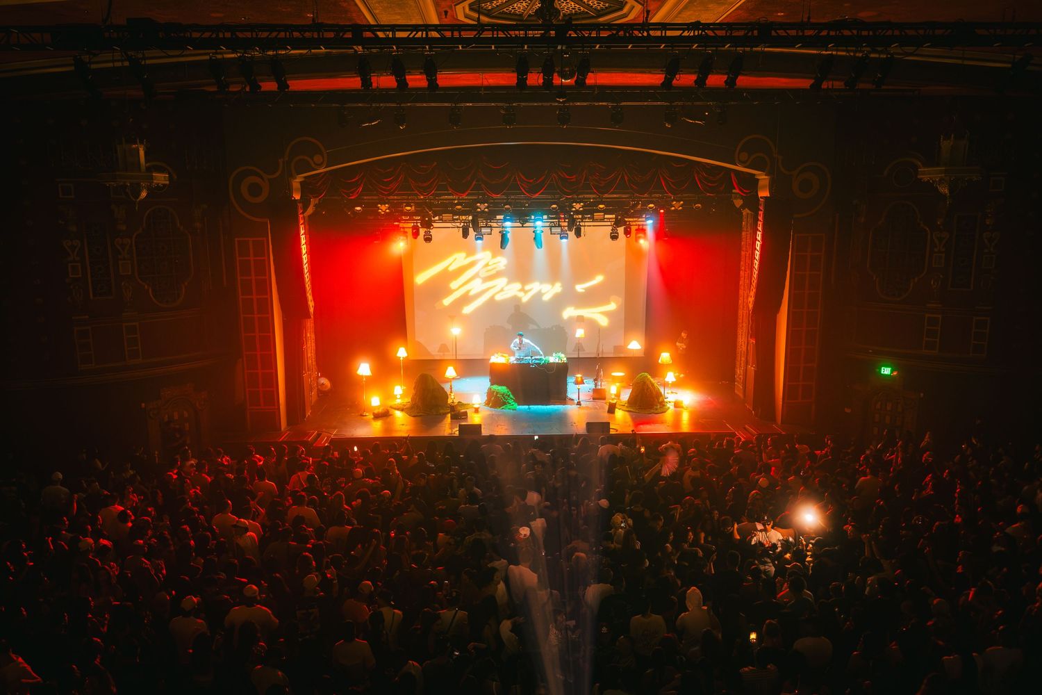 Packed concert hall with red stage lighting, live performers on stage, and an ornate theater venue filled with an enthusiastic crowd.