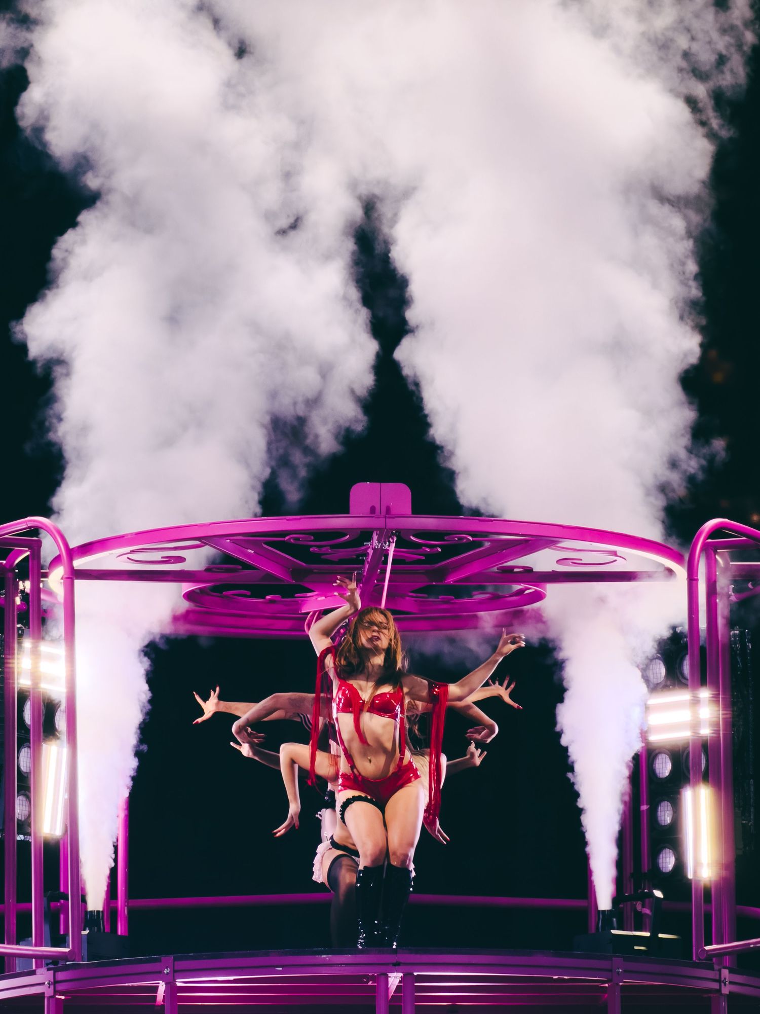 Performer in red lingerie on illuminated pink carousel stage with dramatic smoke effects and bright lights at night.