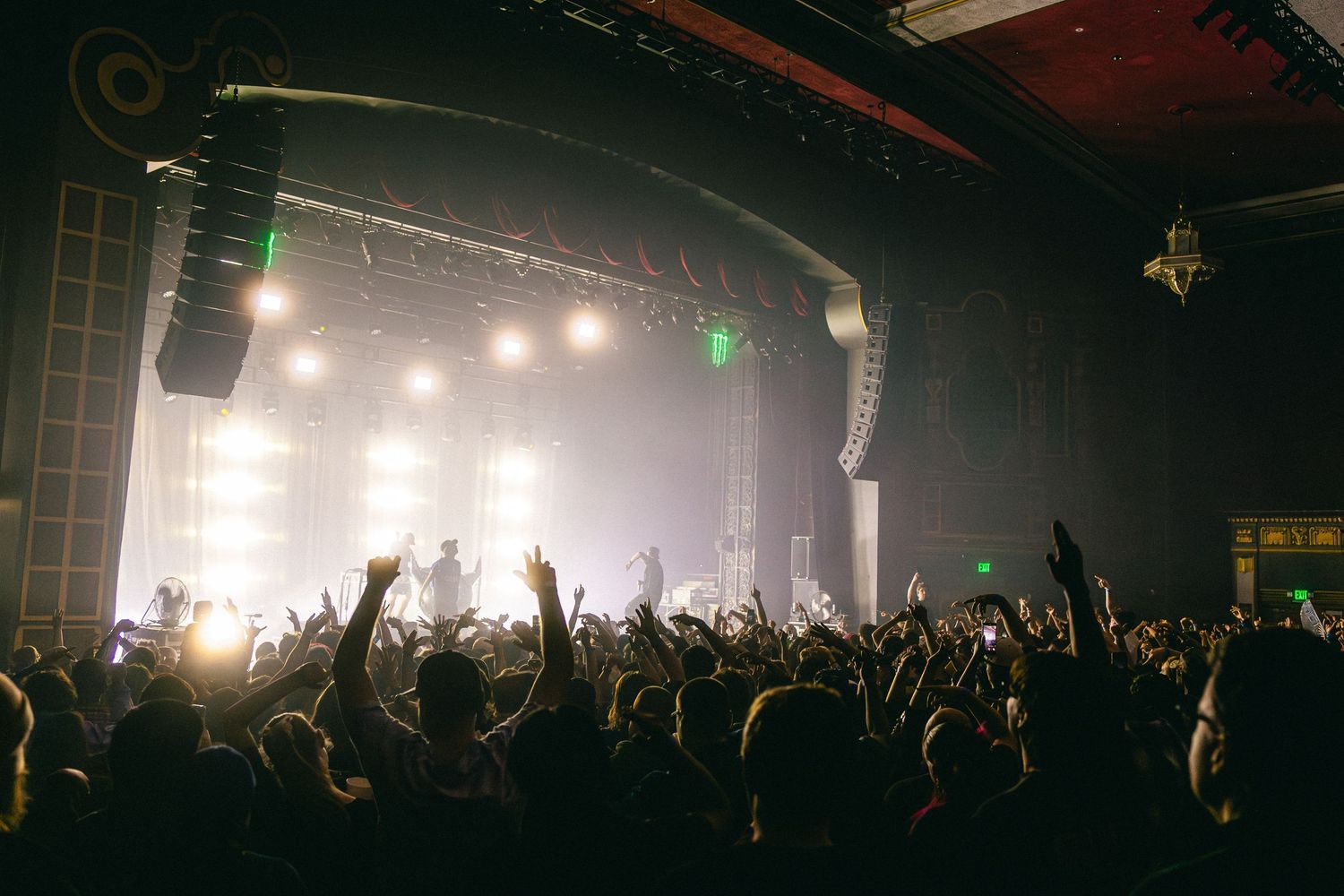 Silhouetted crowd with raised hands at a dark concert venue with bright stage lights creating an atmospheric ambiance.