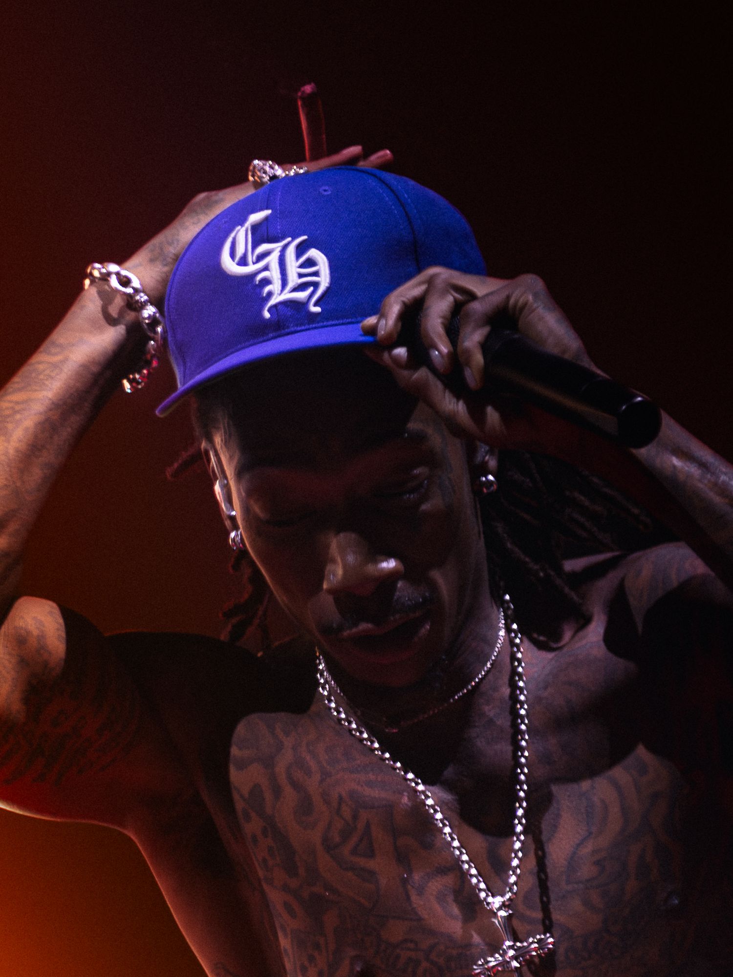 Heavily tattooed shirtless male performer holding microphone and adjusting blue Chrome Hearts snapback cap on stage.