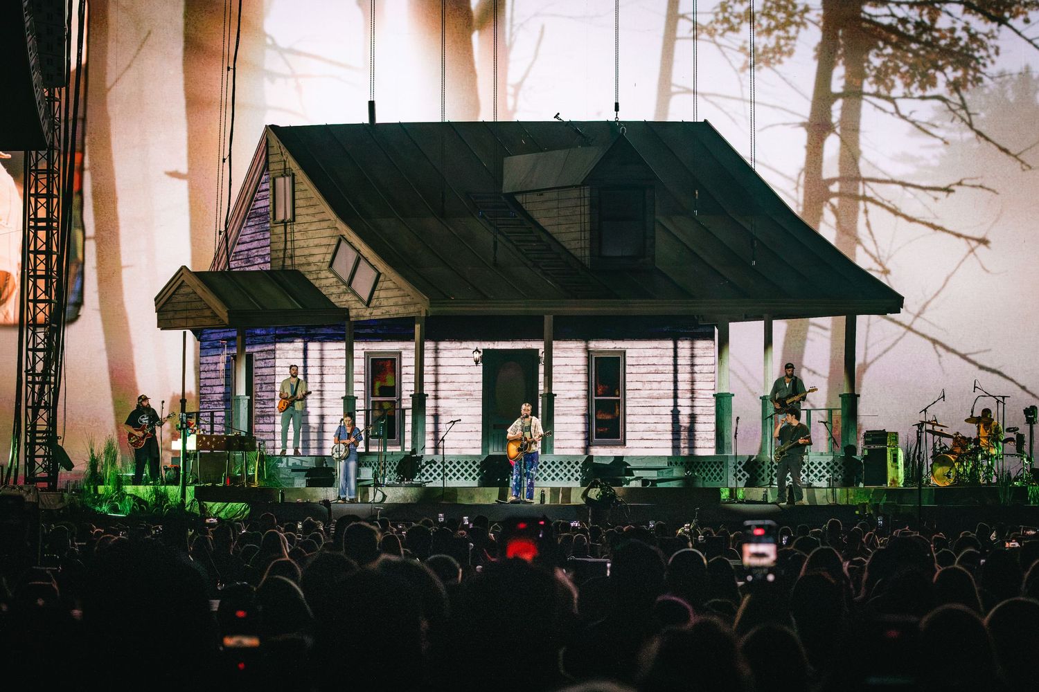 Live concert stage designed as a rustic house with band performing before a large crowd in a misty forest setting.