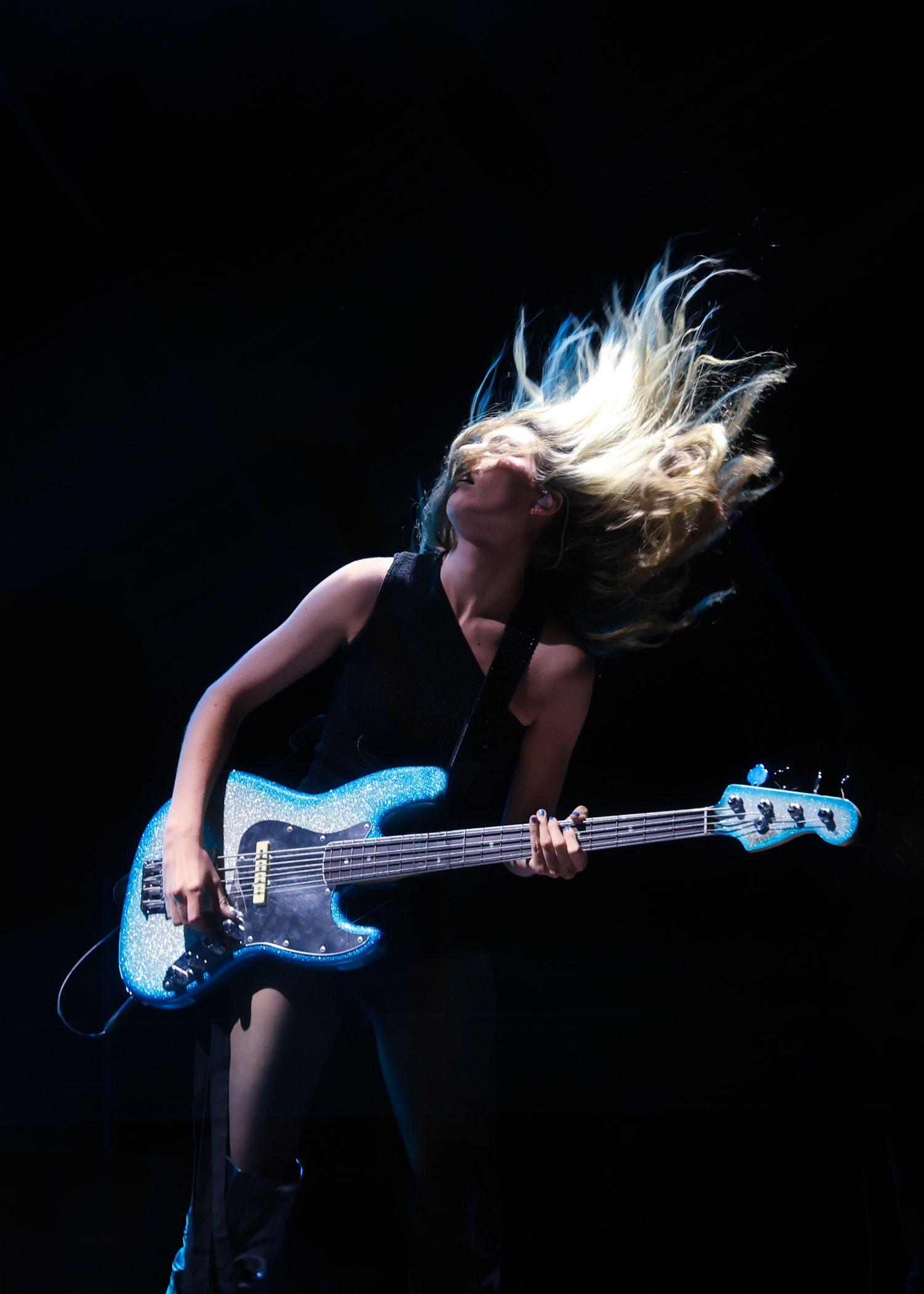 Musician playing blue electric bass guitar with dramatic hair movement on dark stage.