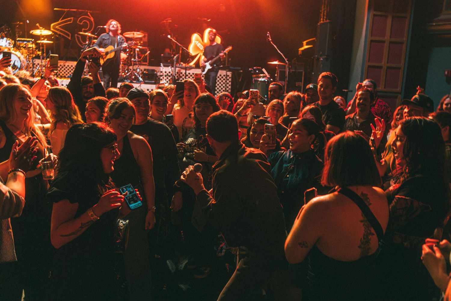 Crowded concert venue with live band performing on stage under warm red and orange lights.