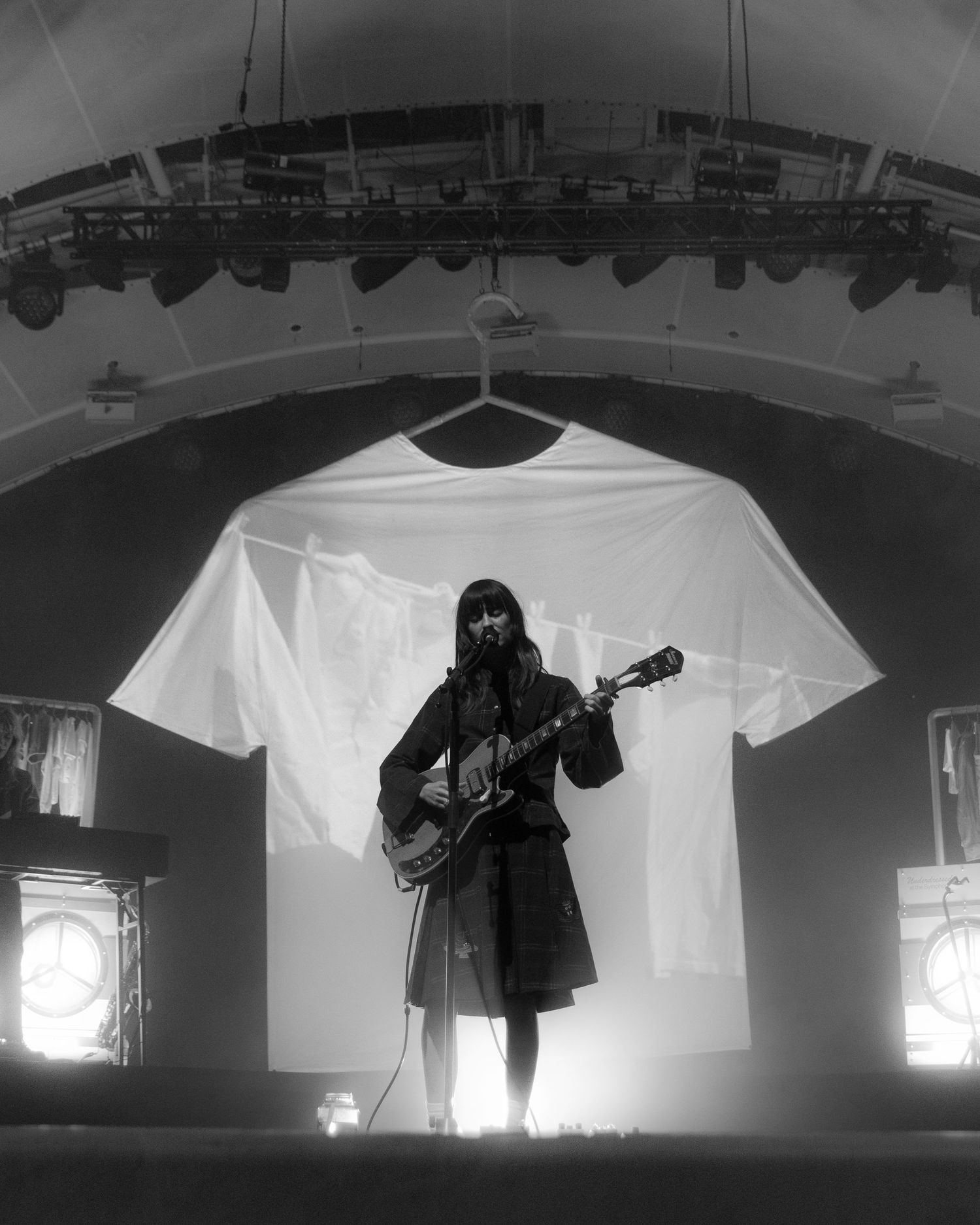 Black and white concert stage with white t-shirts hanging as backdrop and performers in silhouette.