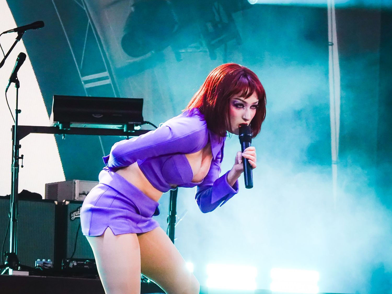 Red-haired female singer performing on stage in purple outfit, holding microphone with dramatic fog lighting.