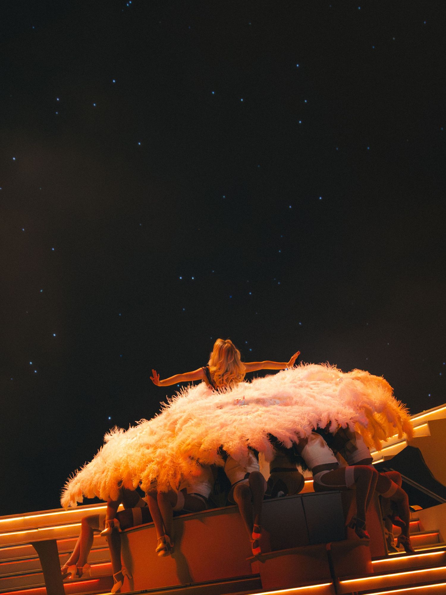 Performer in fluffy pink costume atop a glowing wooden structure under a starry night sky.