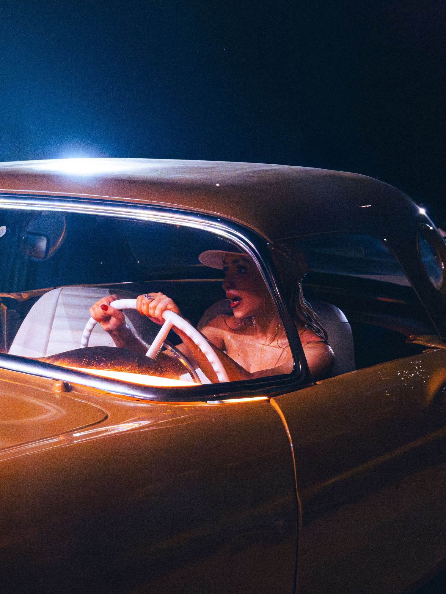 Woman in white dress driving a vintage golden car at night, illuminated by dramatic backlighting.