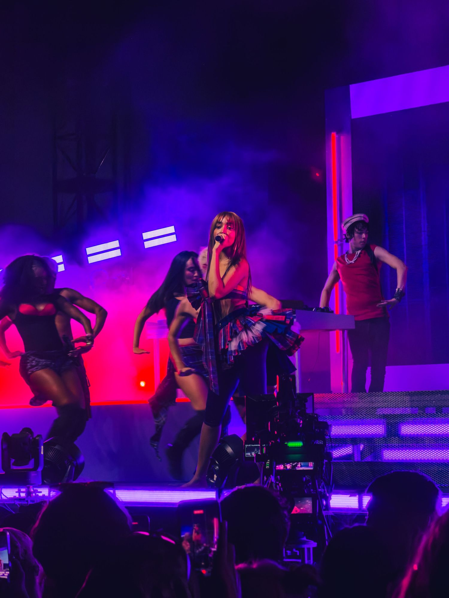 Female pop star performs on stage with dancers under purple and red lights at a live concert.