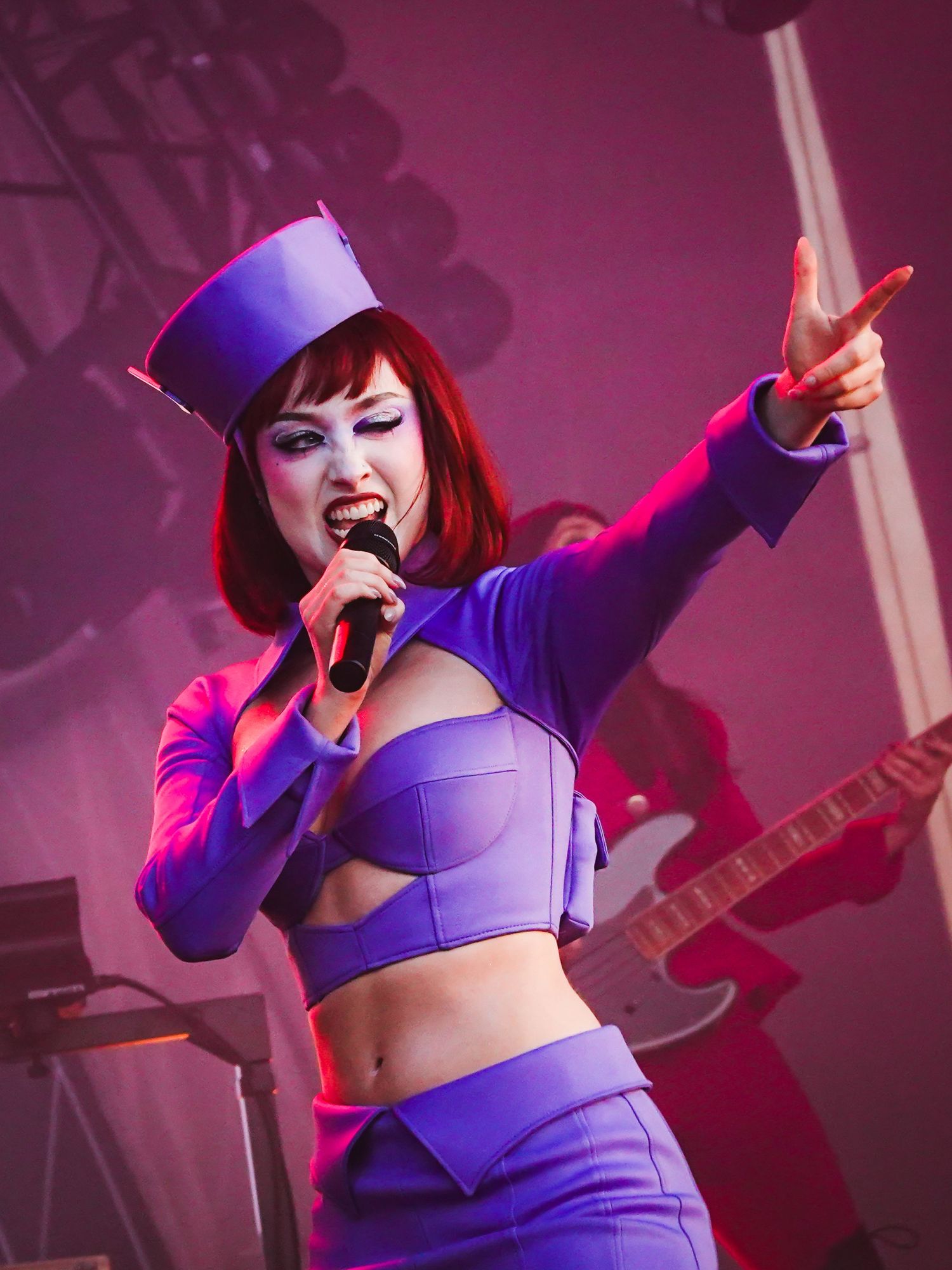 Female singer in purple crop top and hat performing on stage, pointing at audience while holding microphone.