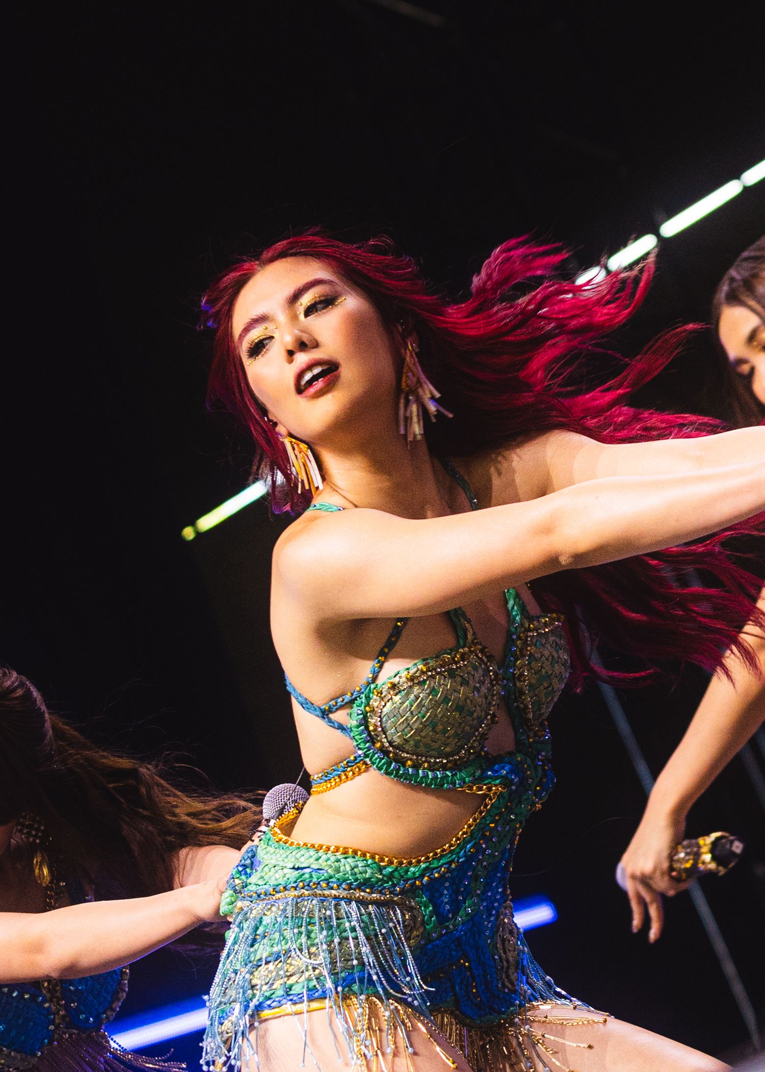 A performer with vibrant red hair dances on stage in a colorful beaded costume under bright lights.