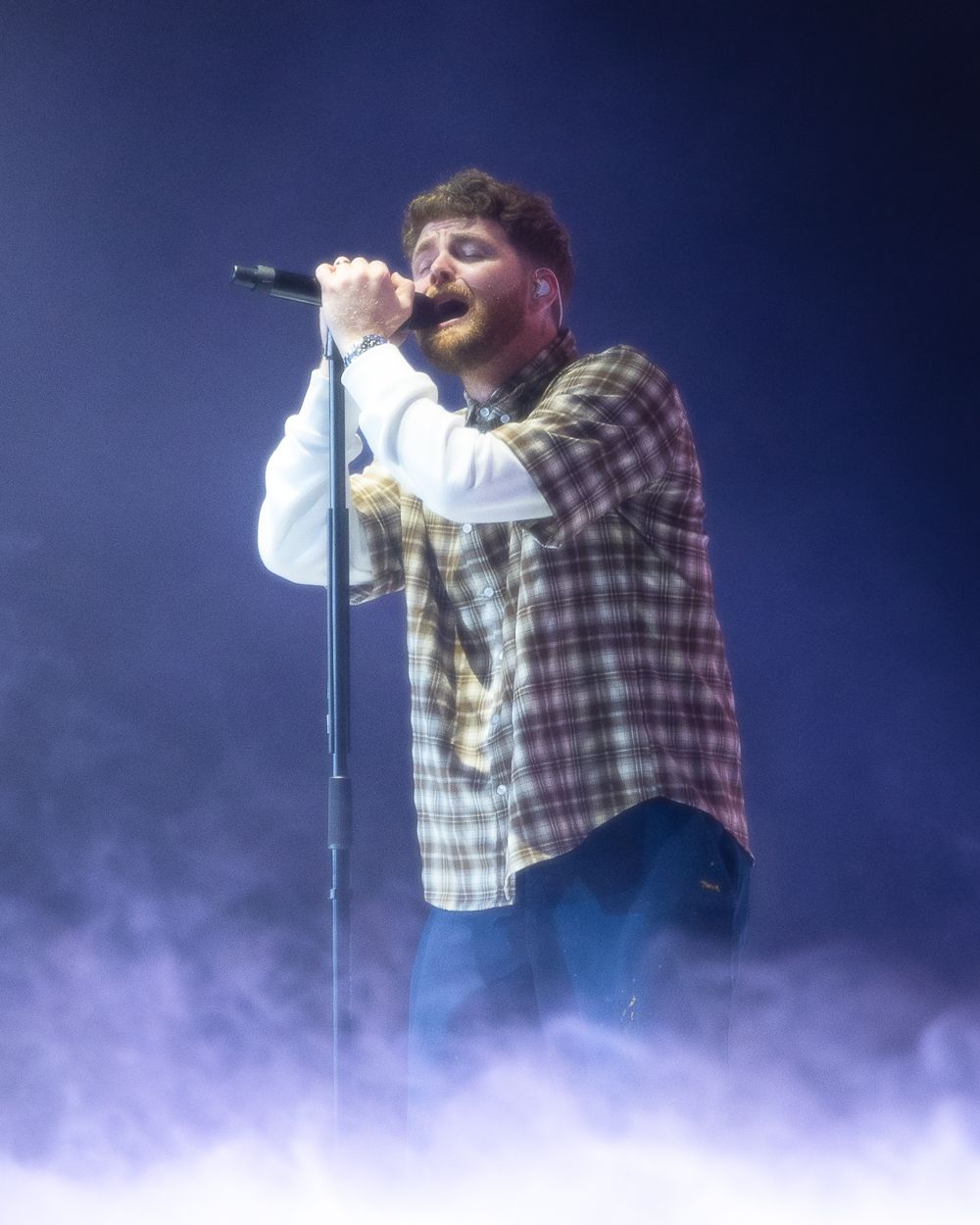 Concert performer singing into microphone on smoky stage wearing plaid flannel shirt.