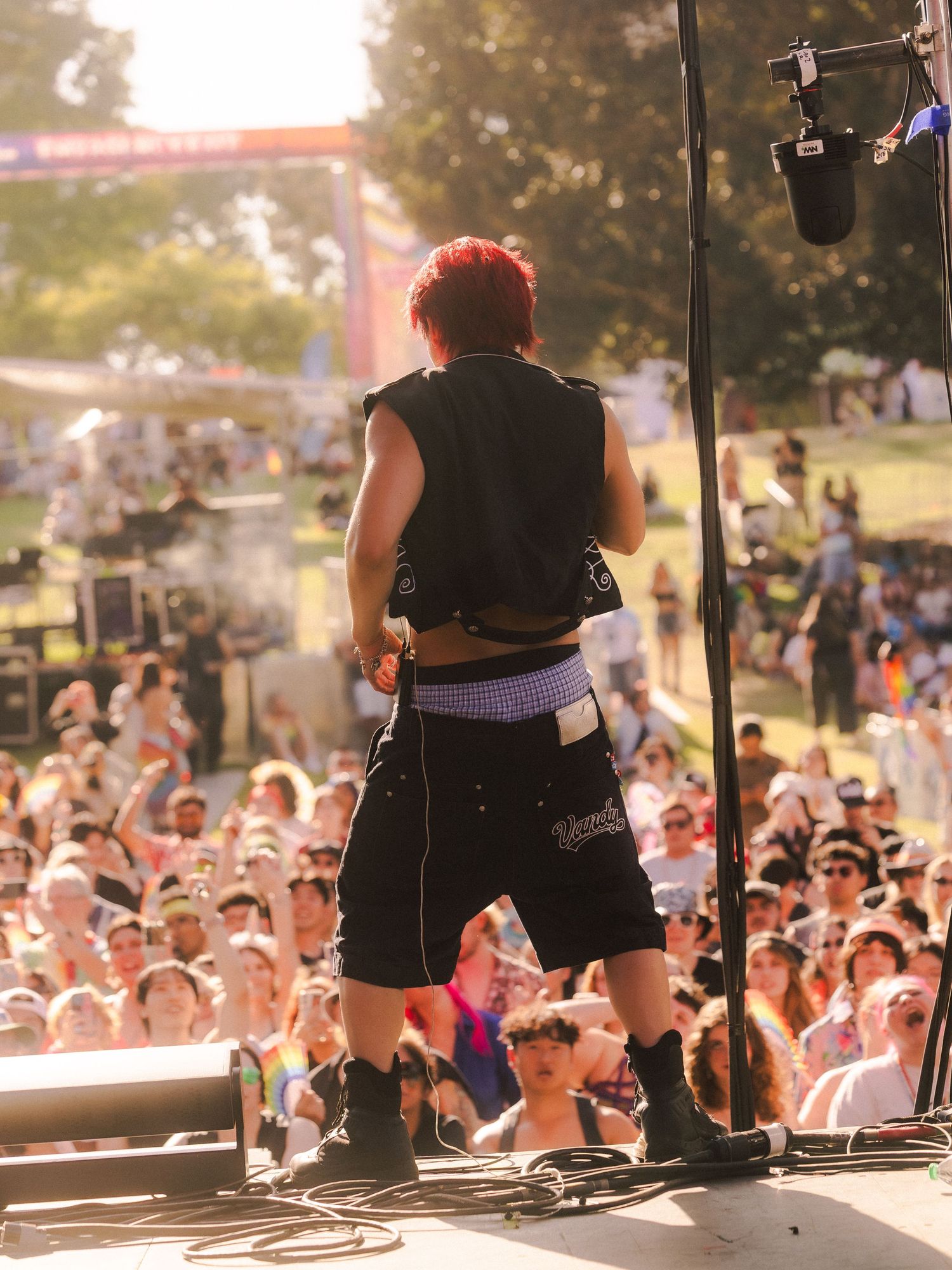 Red-haired punk rock performer on outdoor festival stage with back to crowd, wearing black vest and shorts, boots, purple belt.