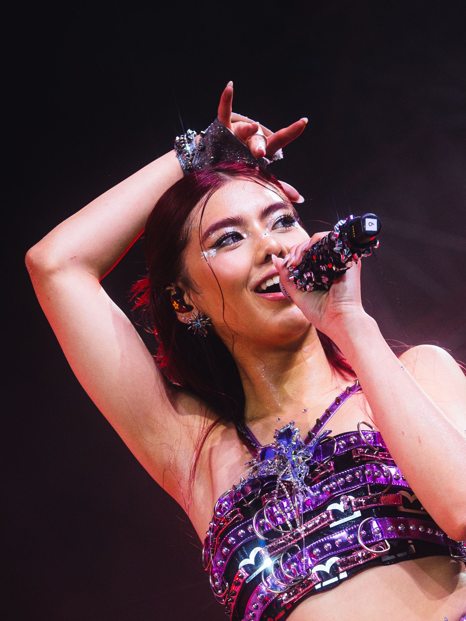 A female performer sings on stage wearing a purple sequined crop top and decorative crown under dramatic concert lighting.