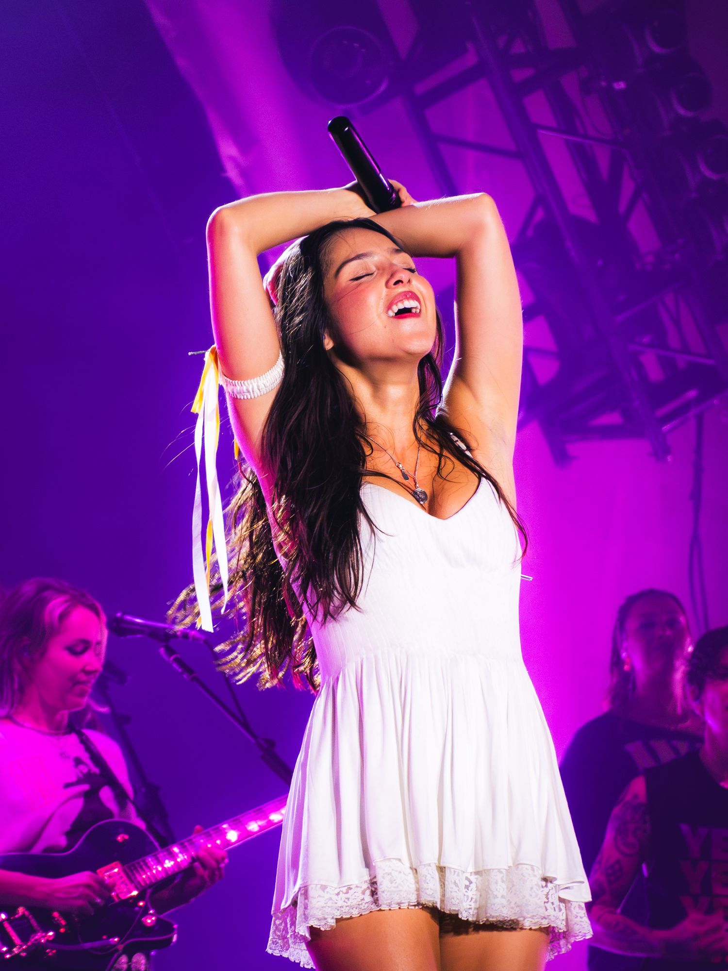 Woman in white dress singing passionately on stage with purple lighting and band members in background.